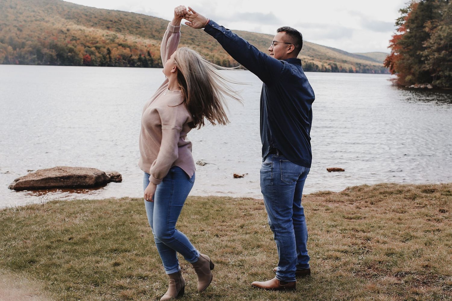 Two people dance together by a scenic lake with mountains and autumn trees in the background.