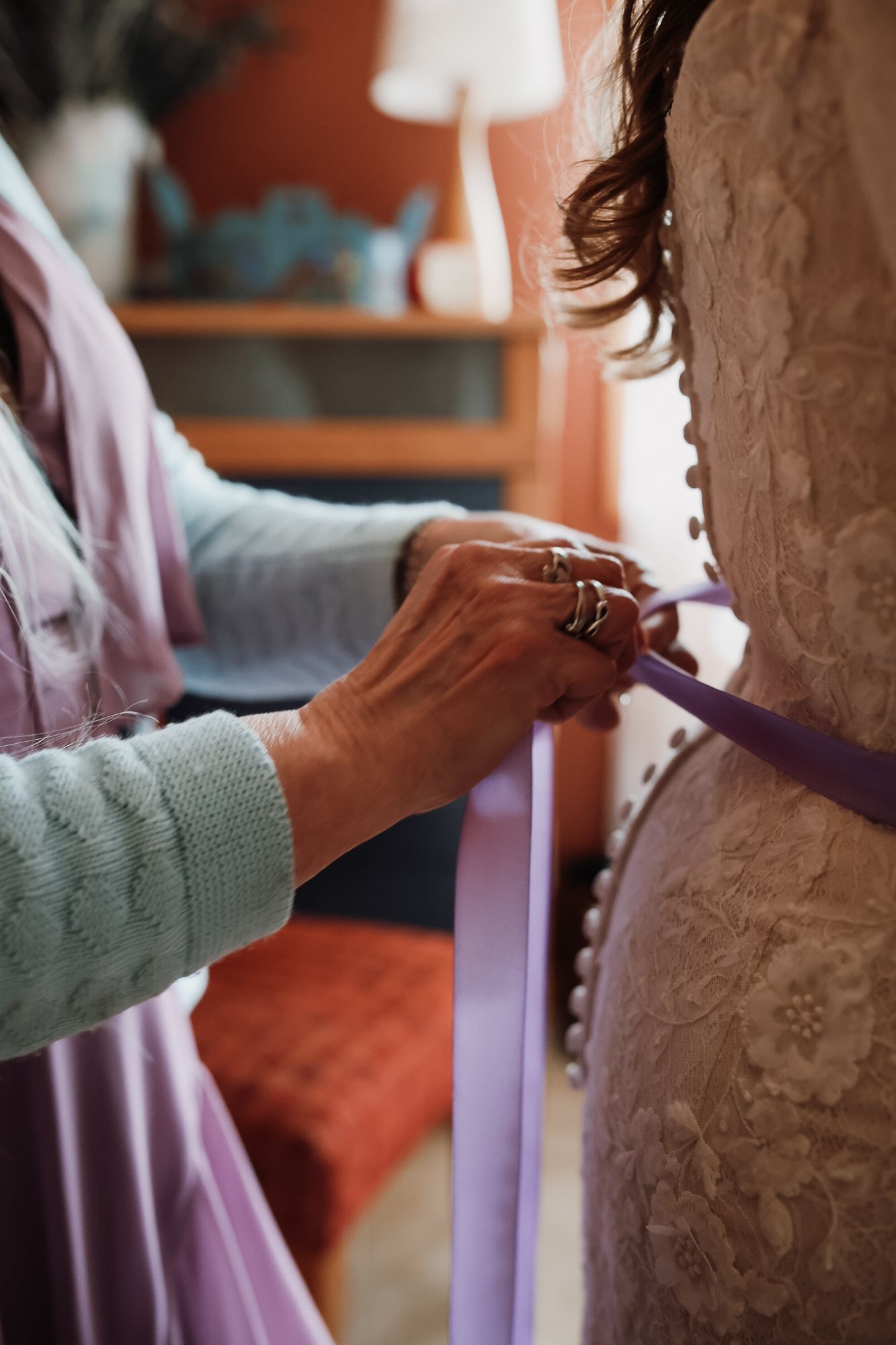 An older woman with rings tying a lavender ribbon on the back of a lace wedding dress indoors.