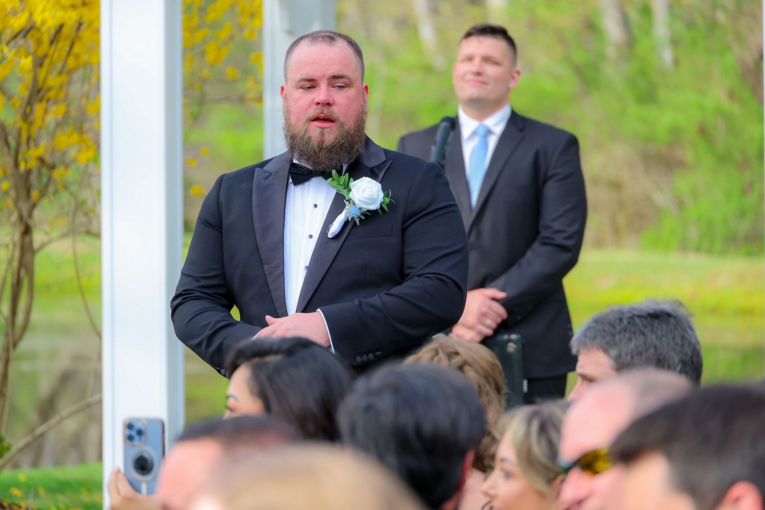 Groom in black tuxedo with white boutonniere watches emotional wedding ceremony outdoors with guests seated.
