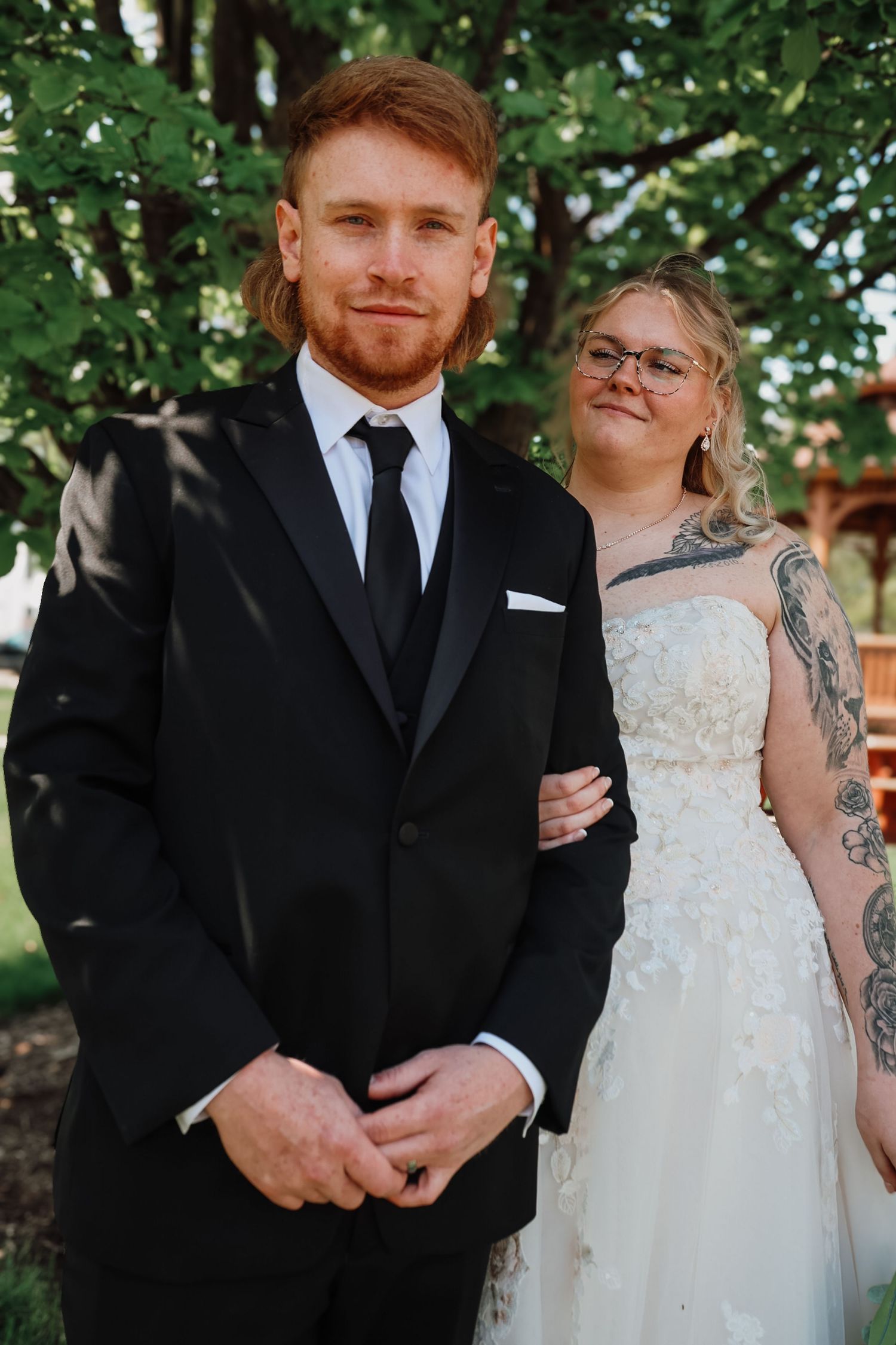 A groom in a black tuxedo stands with a tattooed bride in a white lace gown outdoors at their wedding.