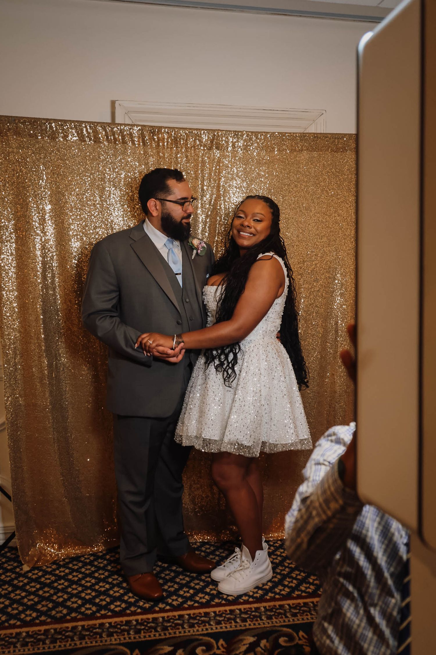 Couple posing together in front of a gold sequin backdrop at a formal event, woman in white mini dress and man in gray suit.