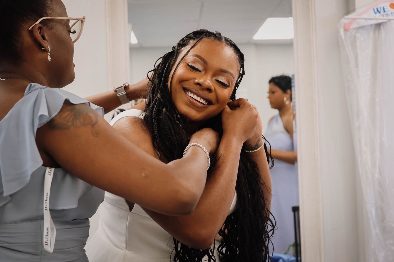 A bridesmaid helps a smiling bride put on a necklace while getting ready for her wedding day.