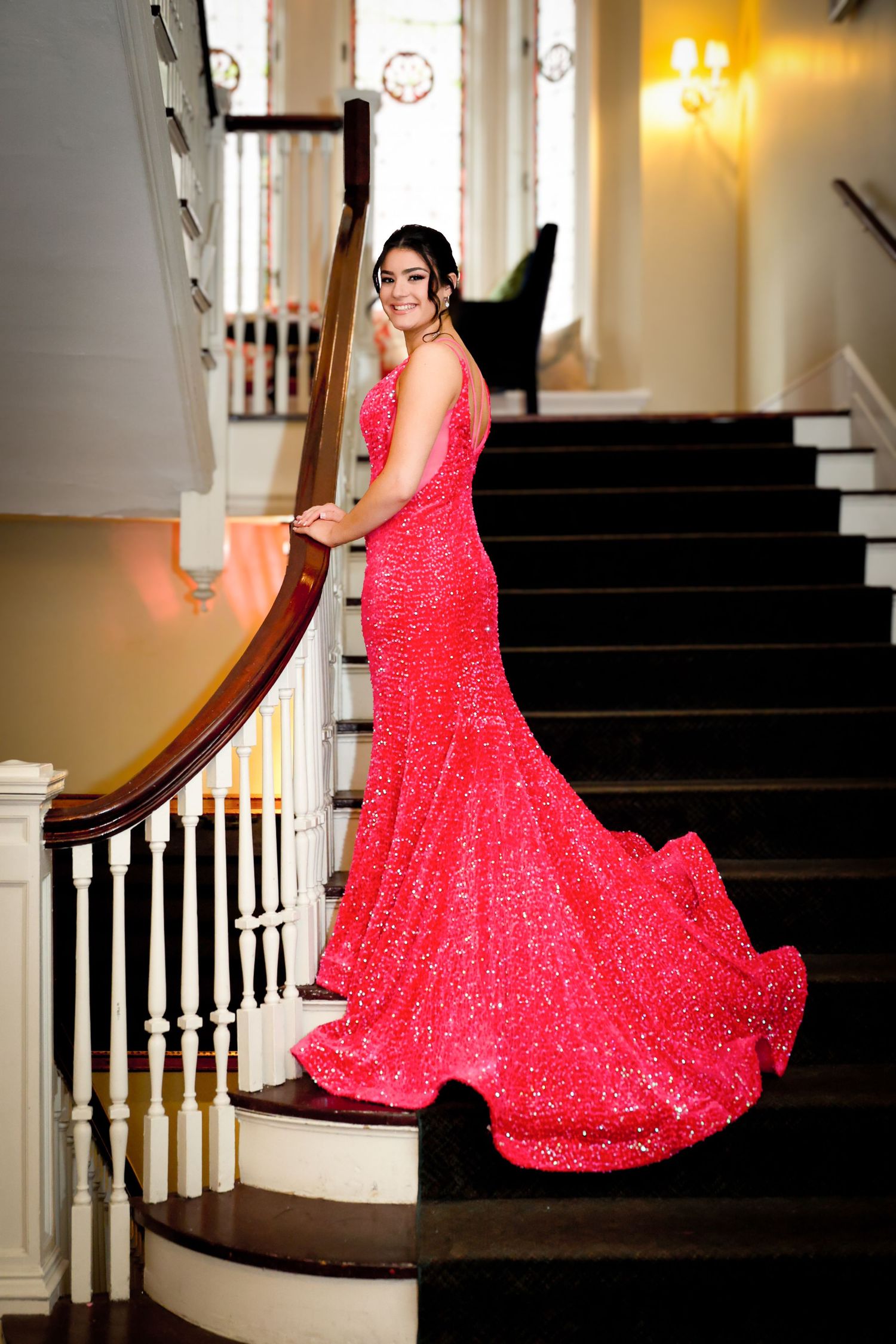 A woman in a sparkling red mermaid gown poses elegantly on a grand staircase with white balusters.