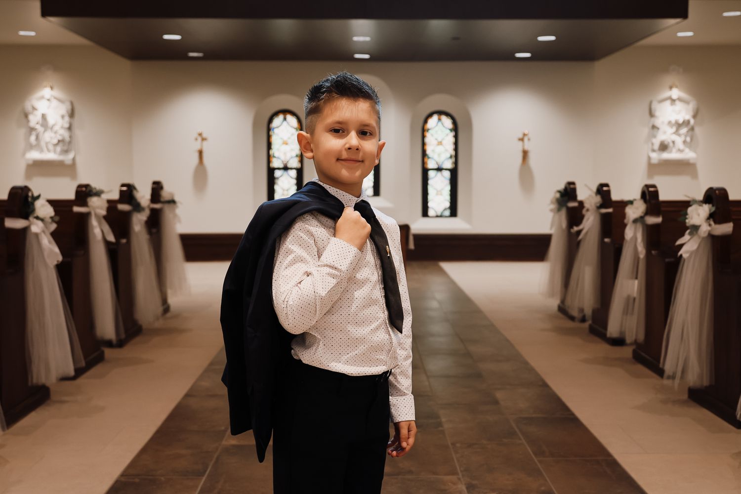 Young boy in formal suit posing in elegant church aisle decorated with floral arrangements for a special ceremony.