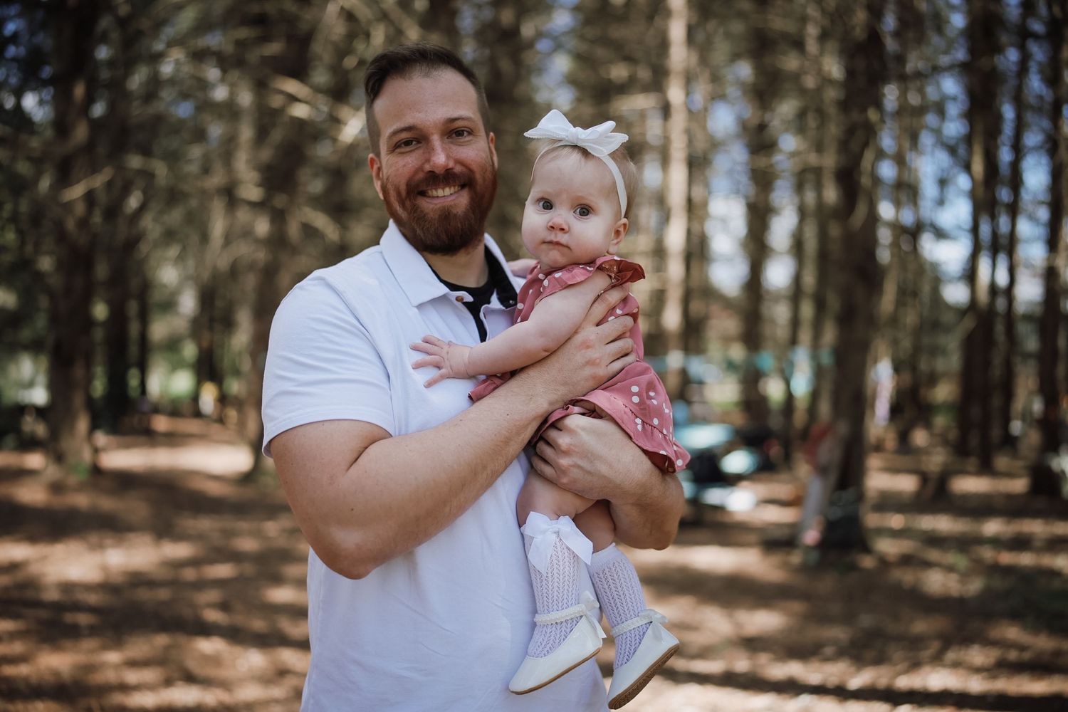 A smiling bearded man in a white polo holds a baby girl in a pink dress with a white bow in a pine forest.