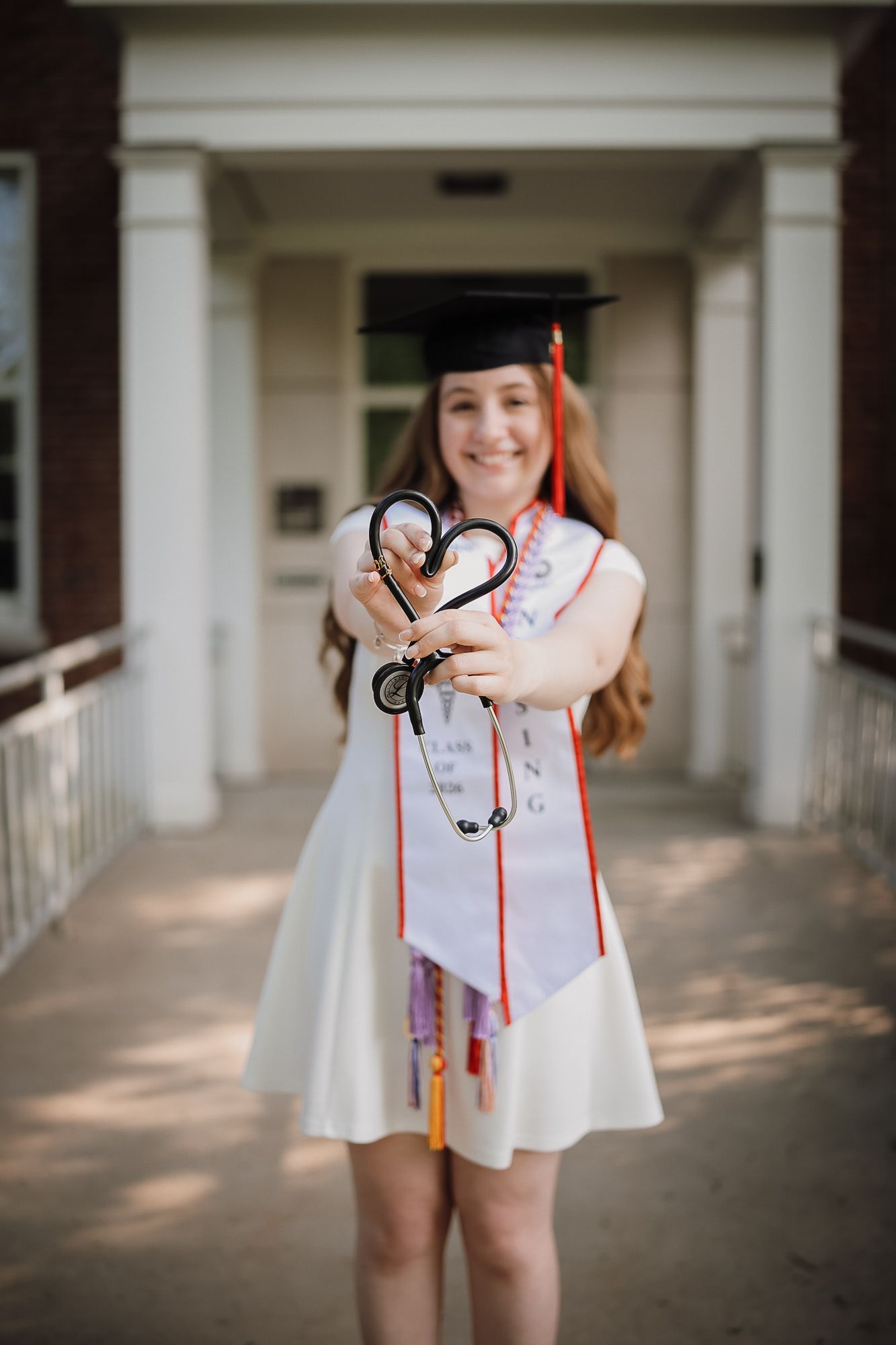 Nursing graduate in cap and gown holds stethoscope in heart shape toward camera outside university building.