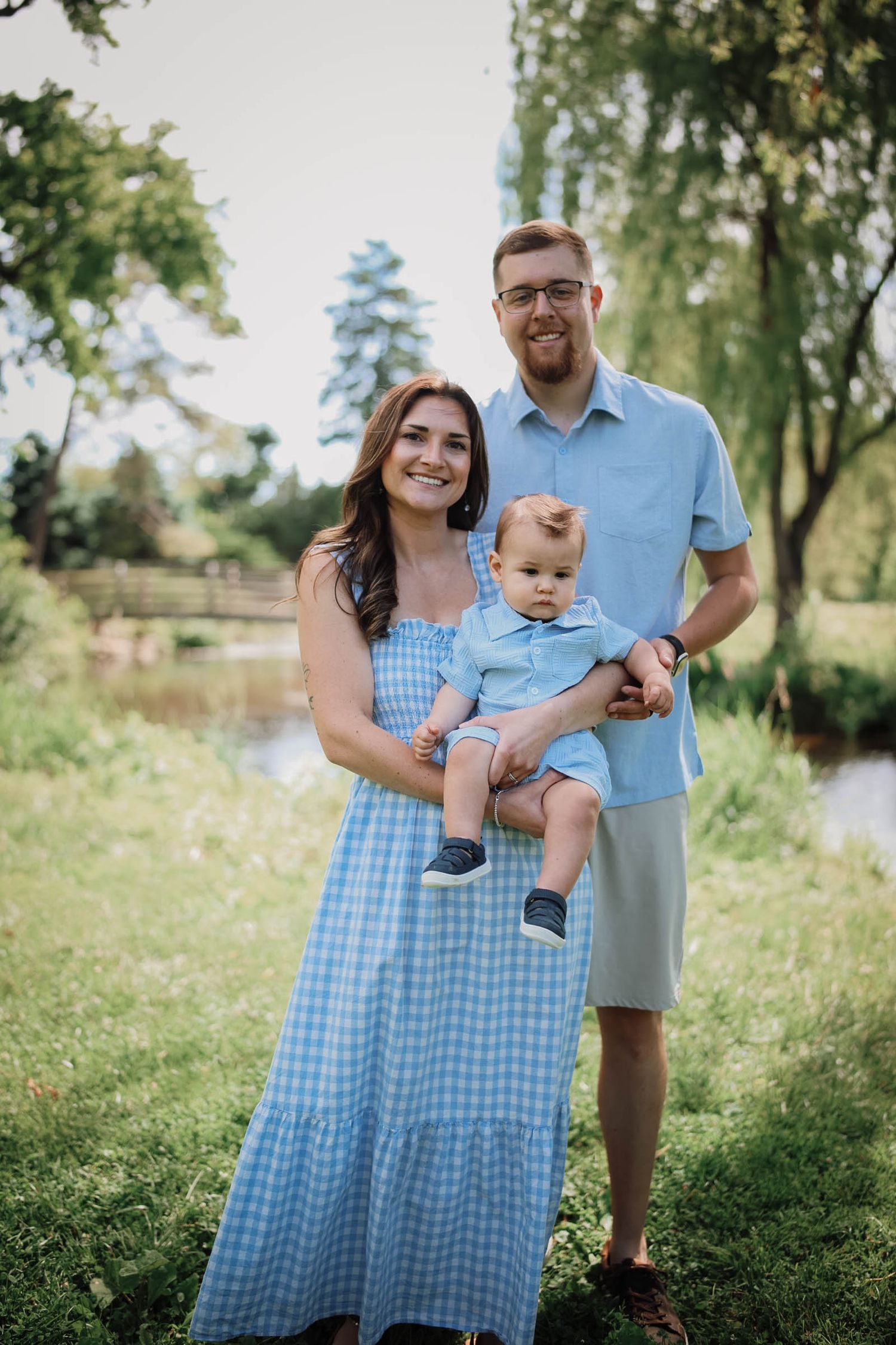 Happy young family with baby boy posing outdoors in matching blue outfits near a scenic park pond.