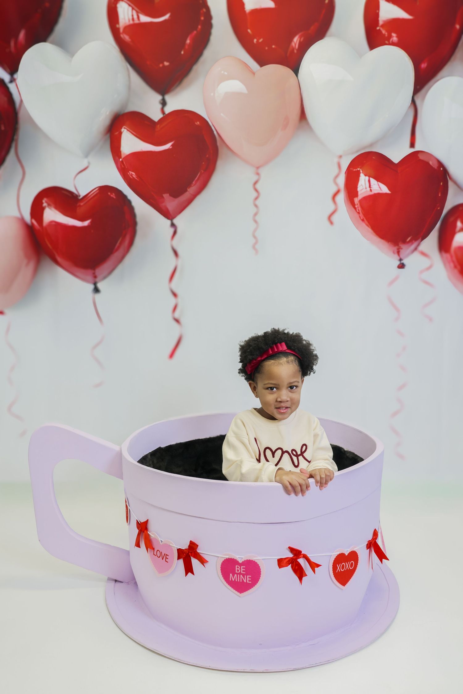 Toddler girl sitting in giant teacup prop surrounded by red, pink, and white heart balloons for Valentine's Day photo.