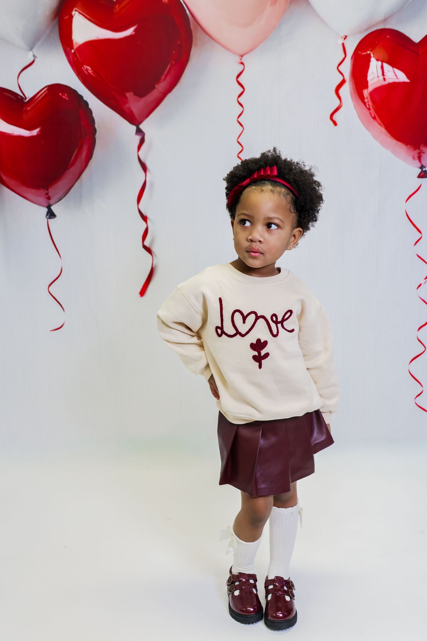 Young girl in cream 'love' sweater and burgundy skirt poses with red heart balloons on Valentine's Day.