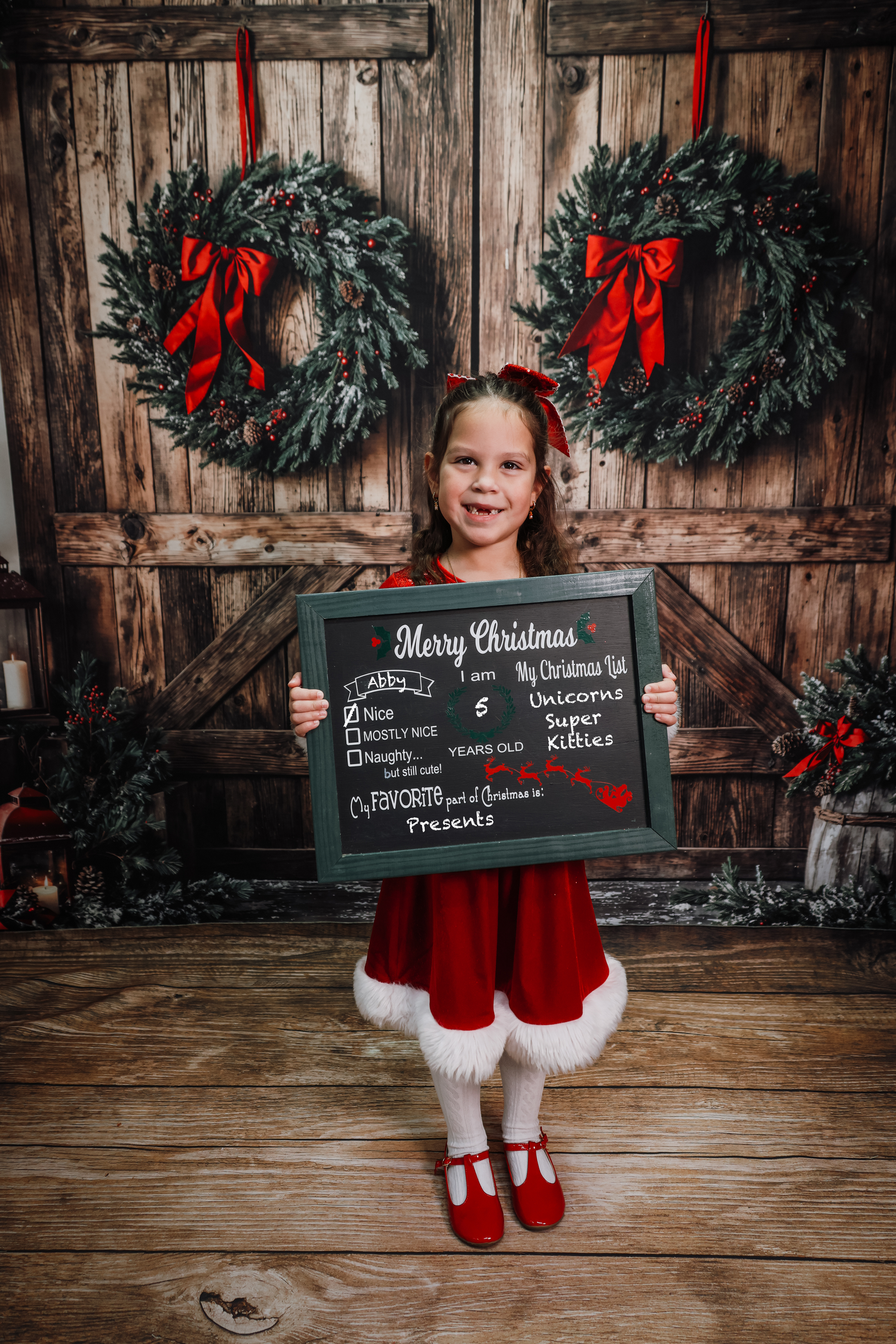 Young girl in red Santa dress holds Christmas chalkboard sign in front of rustic wooden barn doors with holiday wreaths.