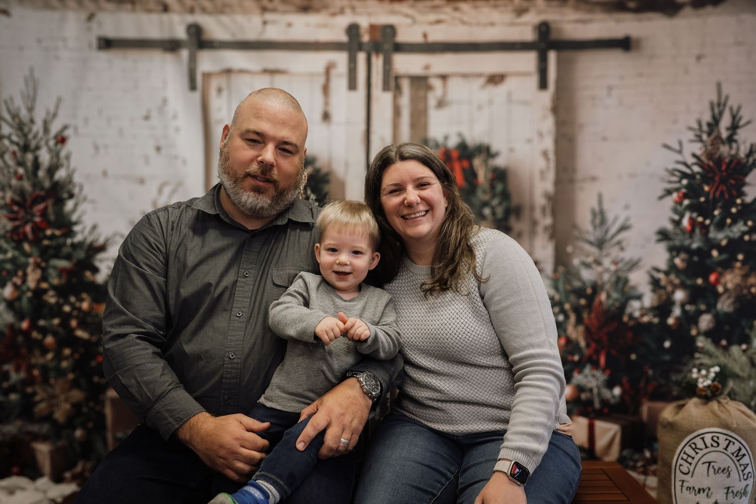 Family of three poses with toddler before decorated Christmas trees in rustic barn setting.