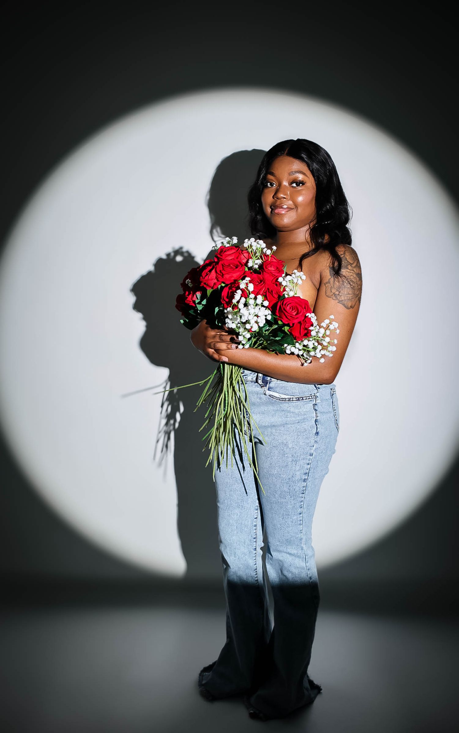 Young woman smiling holding red roses and baby's breath bouquet in jeans under spotlight.