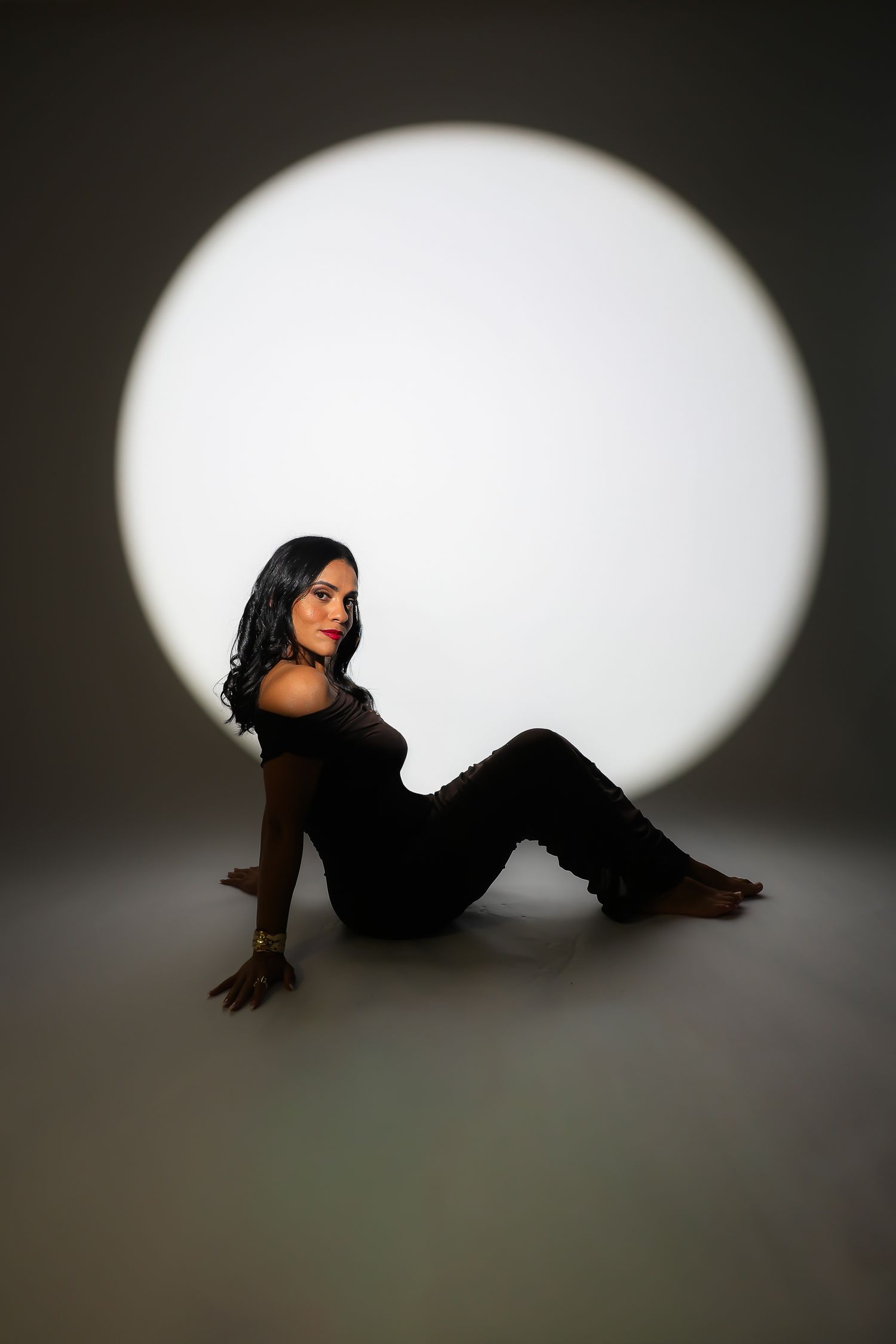Woman in black outfit sitting on floor posed against circular white light backdrop in dark studio.