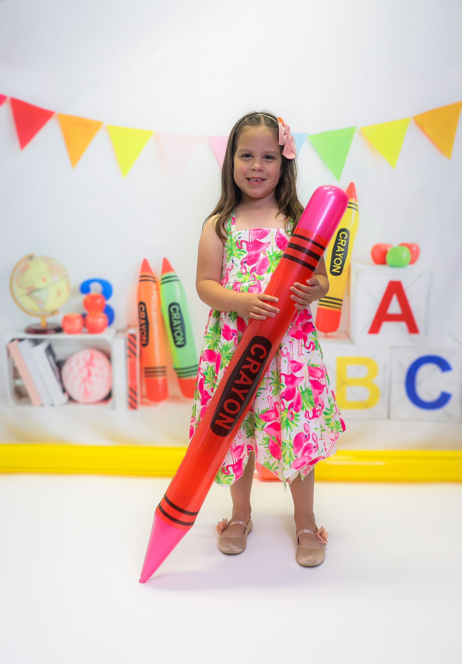 Young girl in floral dress holding giant pink crayon prop in colorful back-to-school themed photo backdrop.