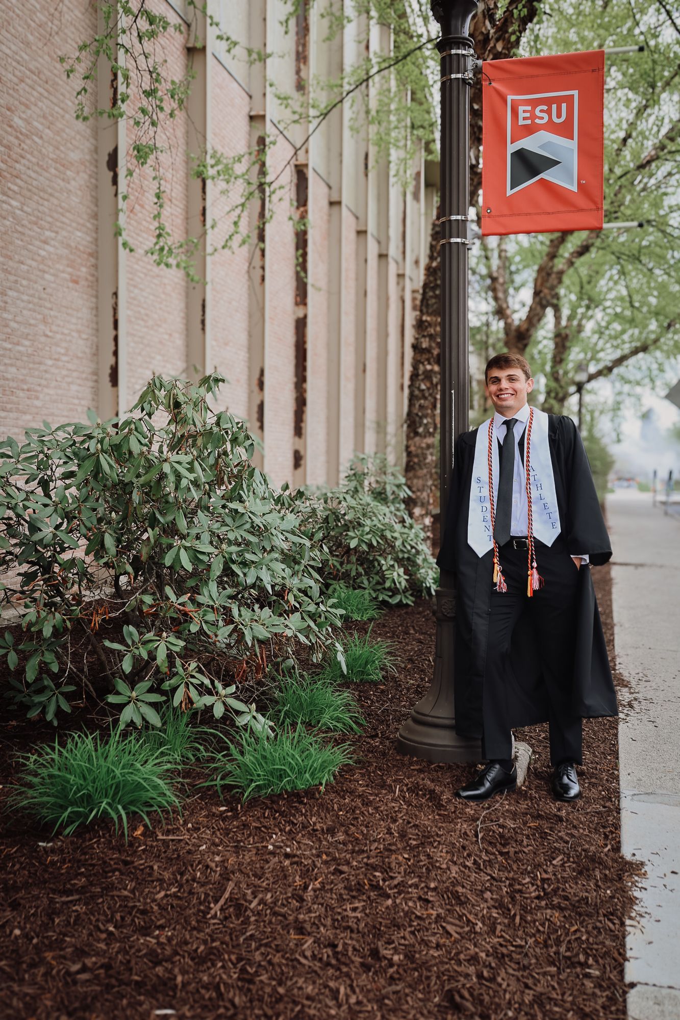 Graduate in cap and gown poses by ESU university banner on campus sidewalk surrounded by lush greenery.