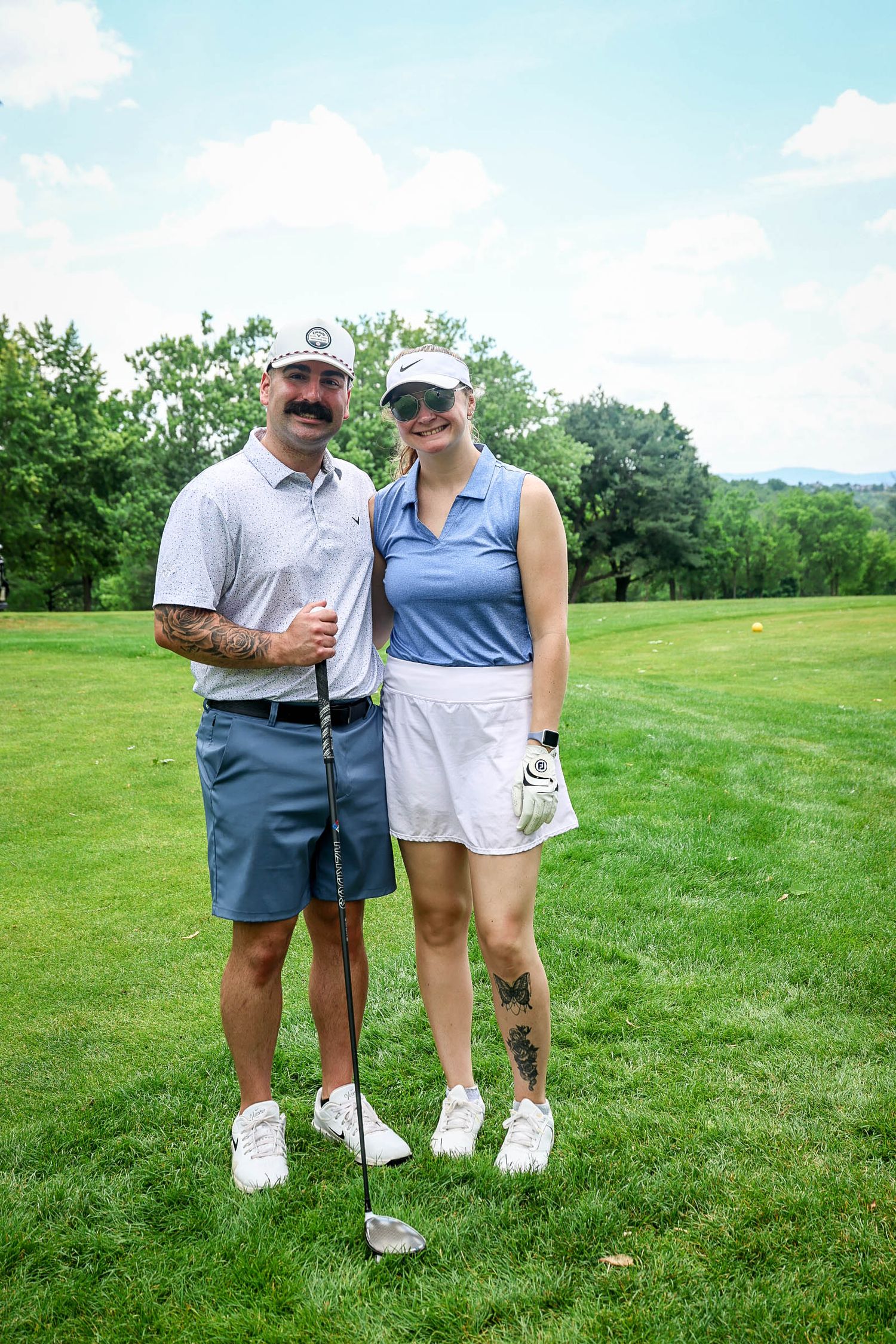 A couple in matching golf attire poses smiling on a lush green golf course on a sunny day.