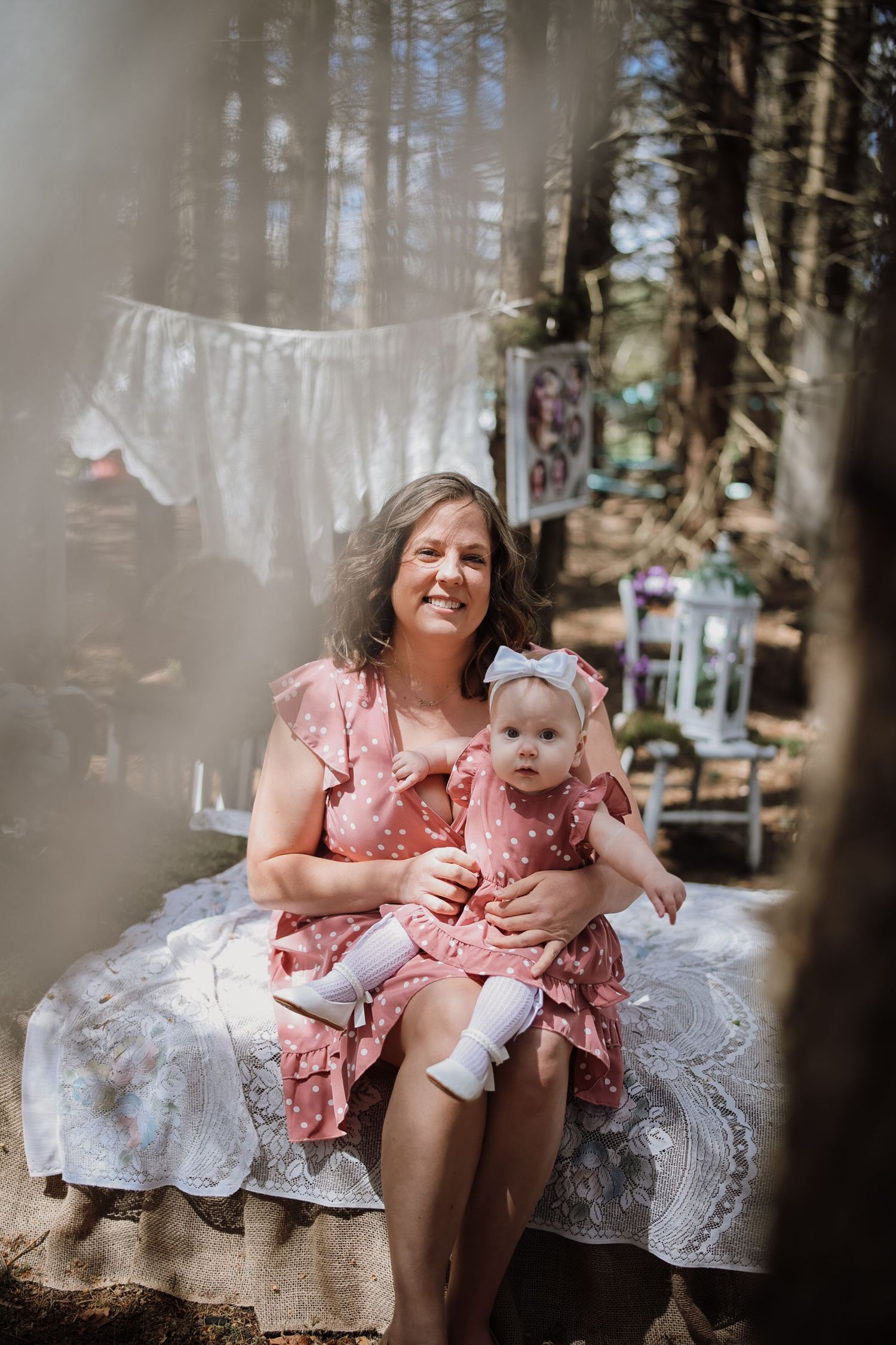 Woman and baby in matching pink polka dot dresses sitting outdoors in a whimsical forest setting.