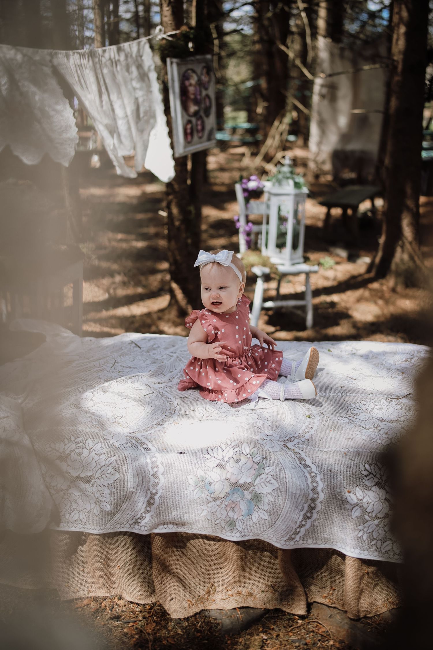 Baby girl in pink polka dot dress sitting on lace blanket outdoors in a whimsical woodland photo shoot.