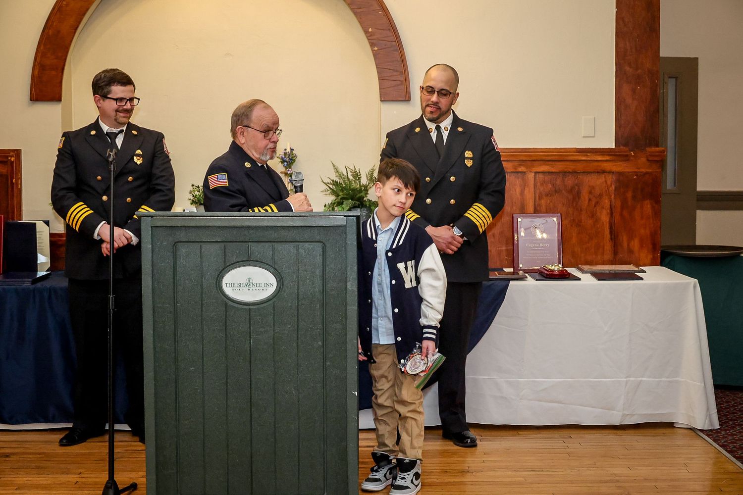 Firefighters in dress uniforms stand at a podium with a young boy during a formal awards ceremony indoors.