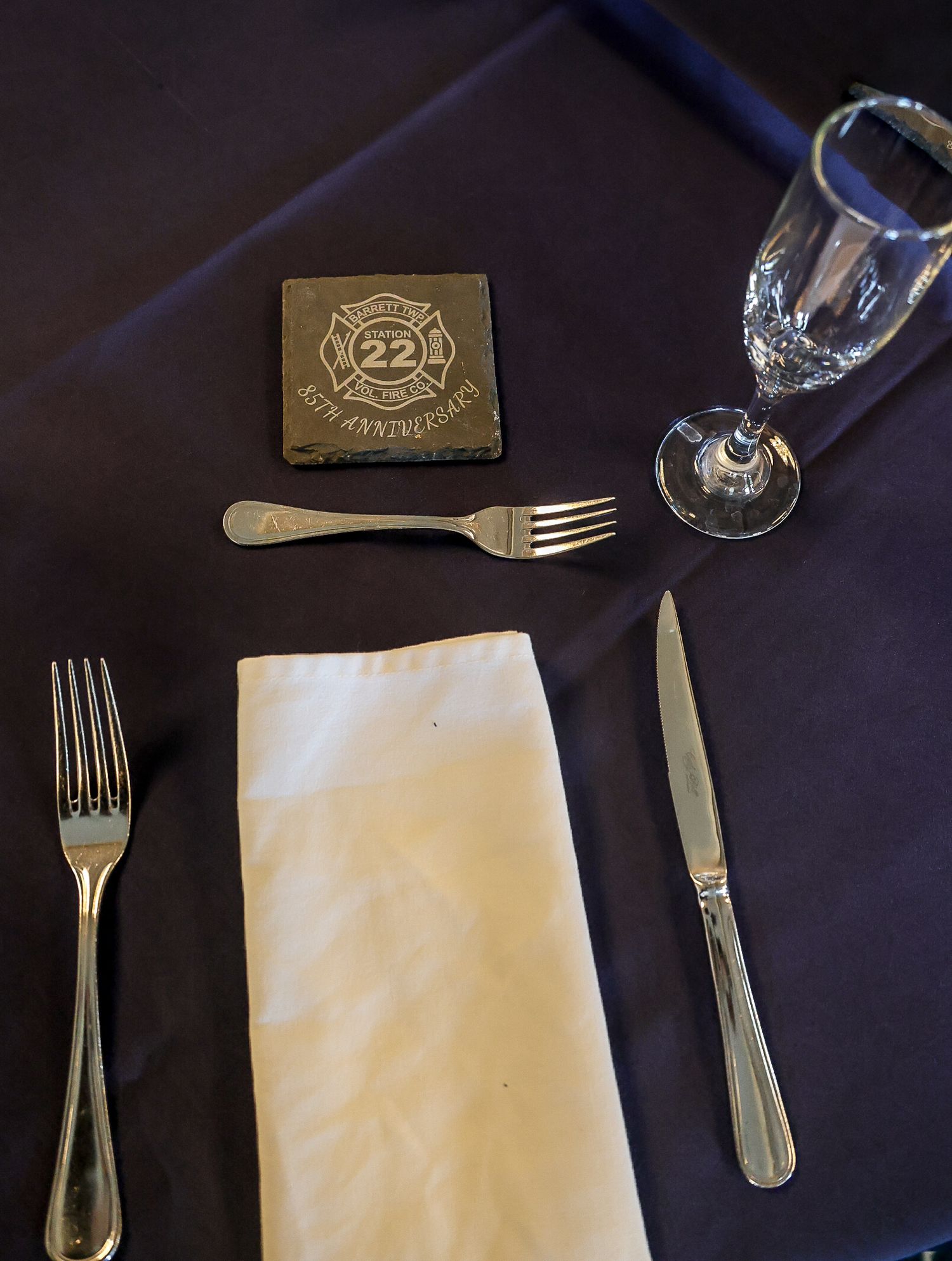 Formal place setting with forks, knife, napkin, crystal glass, and a 22nd anniversary firefighter commemorative coaster.