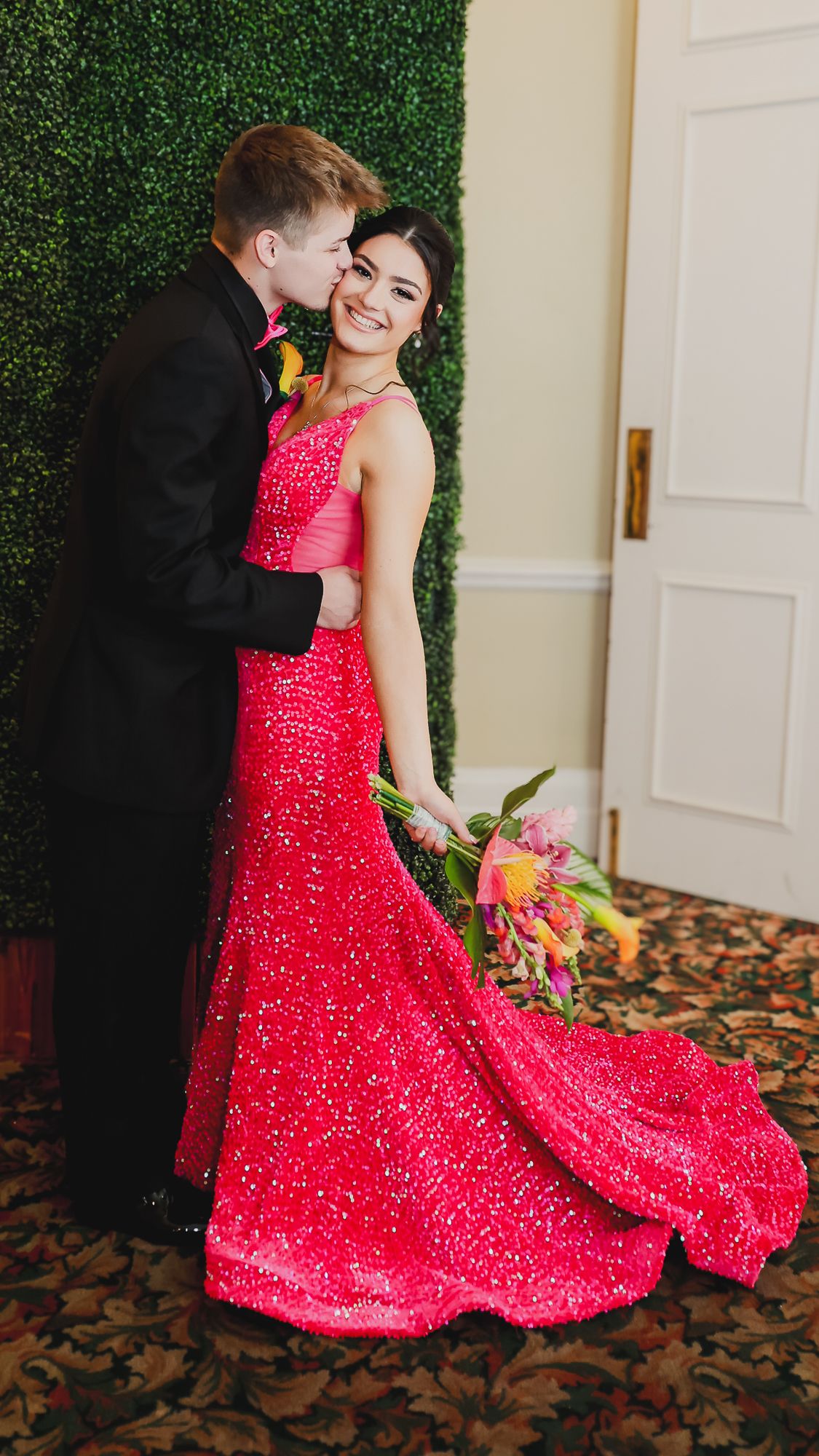 A couple poses for prom, woman in red sequin gown holding flowers while man in black tuxedo kisses her cheek.