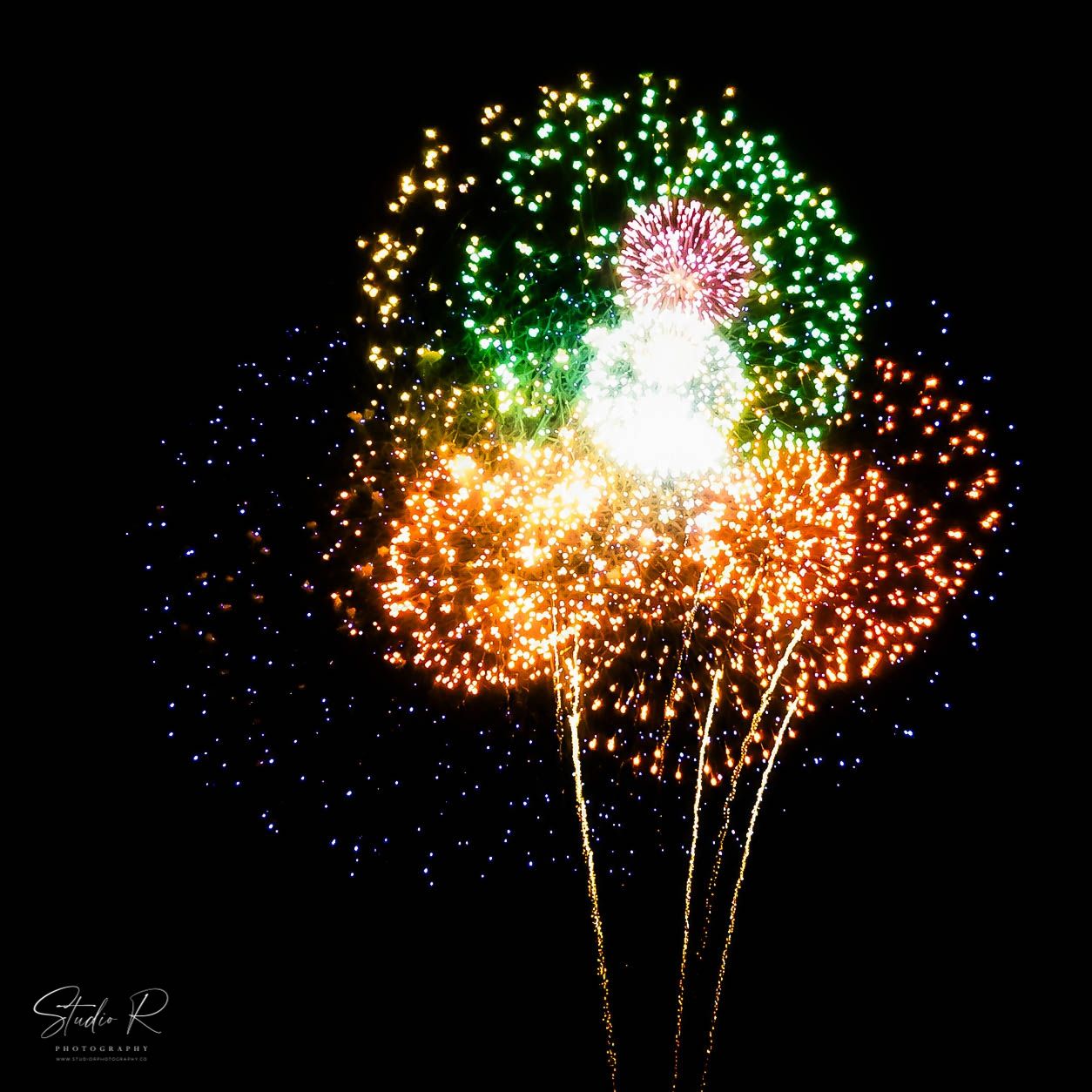 Colorful fireworks burst against a dark night sky, displaying vibrant green, orange, pink, and blue sparks in a stunning display.
