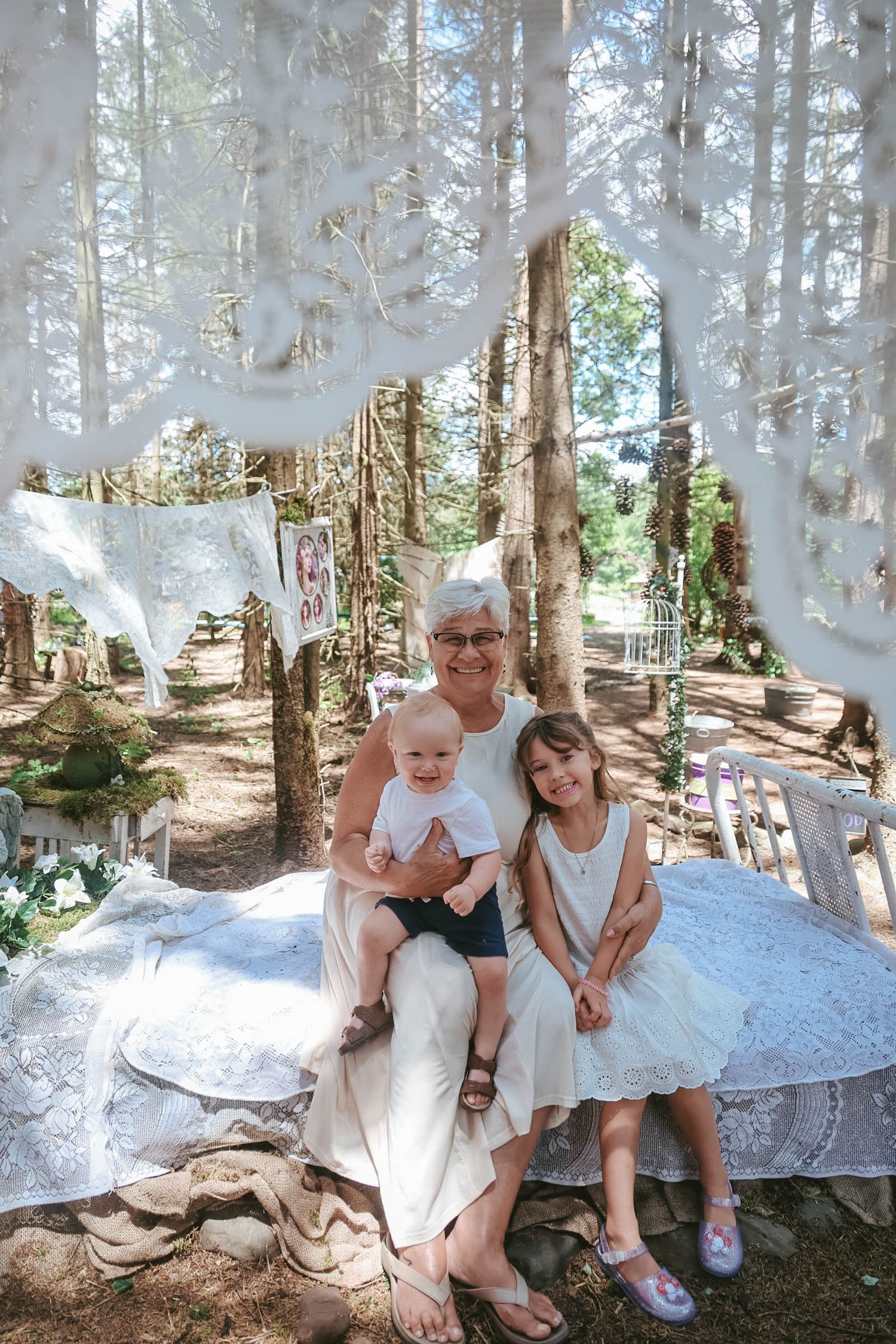 Grandmother sitting outdoors with two grandchildren under white lace decorations in a woodland setting.
