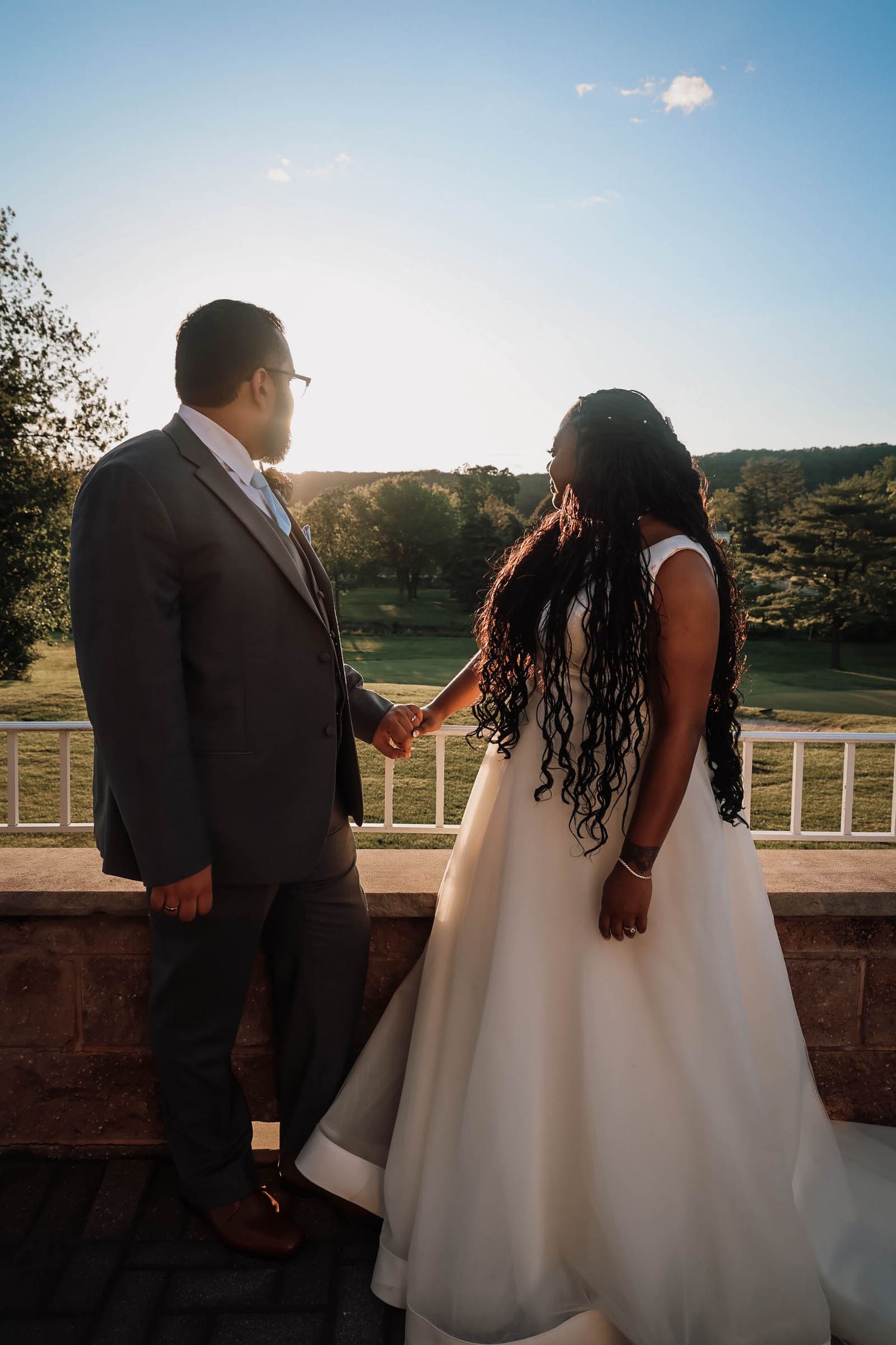 A bride and groom hold hands on a sunlit terrace, overlooking lush green hills during their outdoor wedding ceremony.