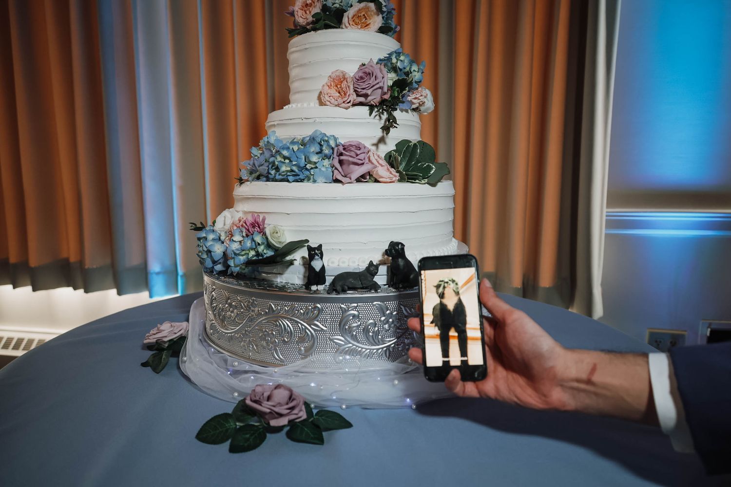 Elegant multi-tier white wedding cake with floral decorations and couple toppers, with a phone displaying the newlyweds.