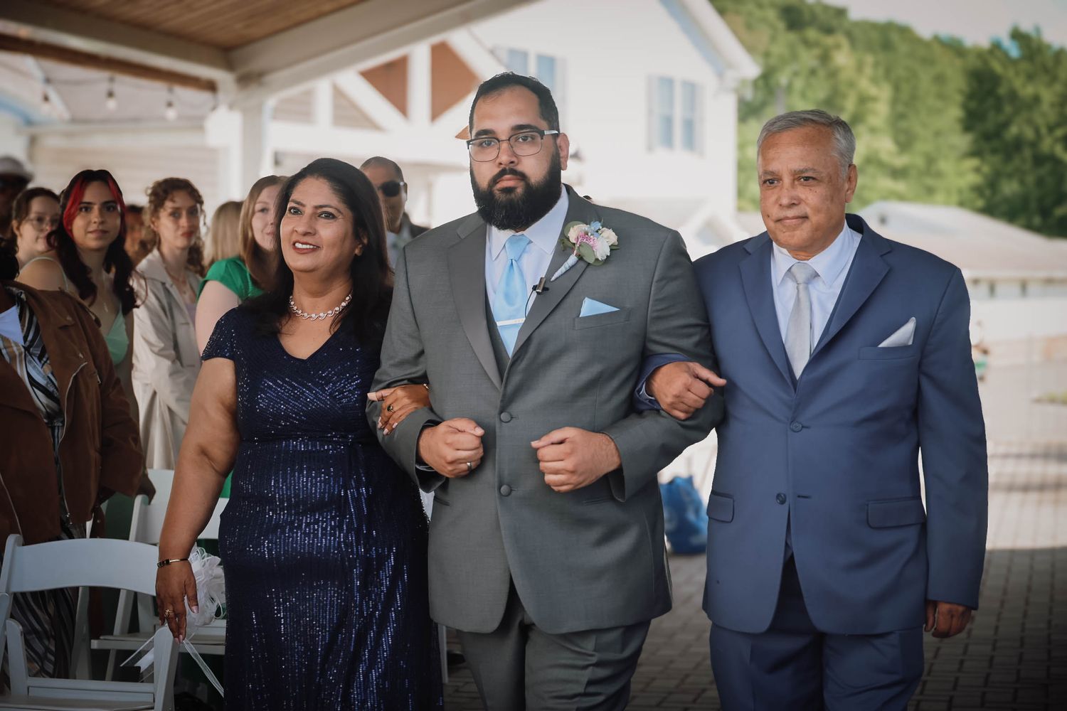 Groom walks down the aisle escorted by his parents at an outdoor wedding ceremony.