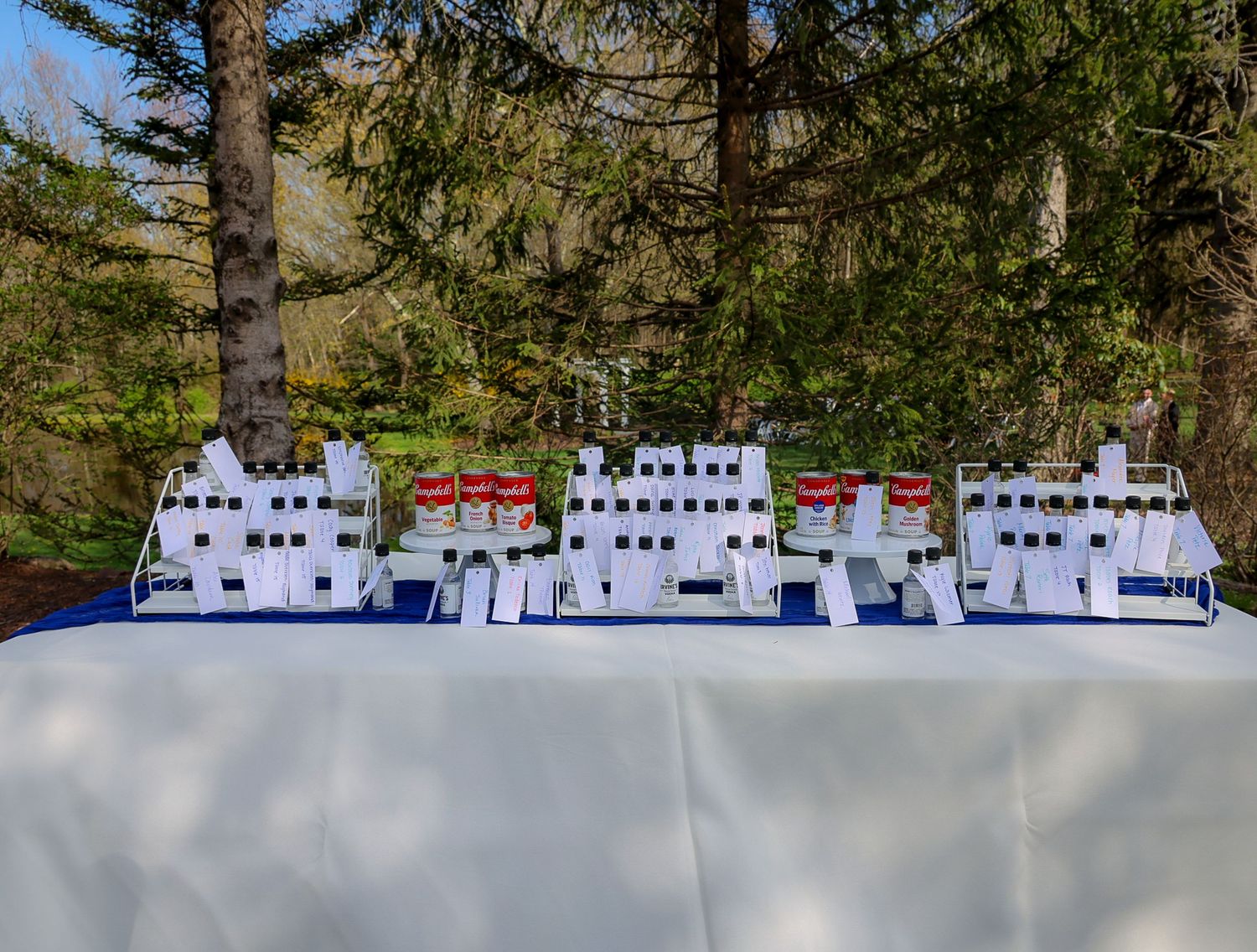 Outdoor display table with white favors and red-labeled items arranged in rows, set against lush green pine trees.