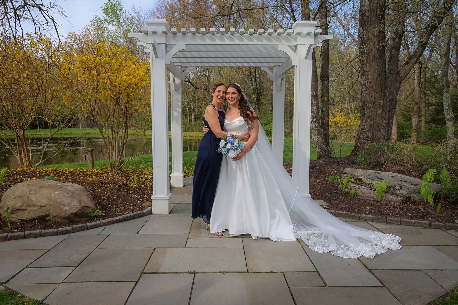 Bride in white gown with bridesmaid in navy dress posing under white pergola in lush garden setting.