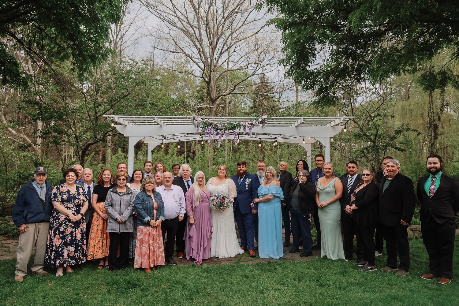 Large wedding party poses together outdoors near a white gazebo surrounded by lush green trees.