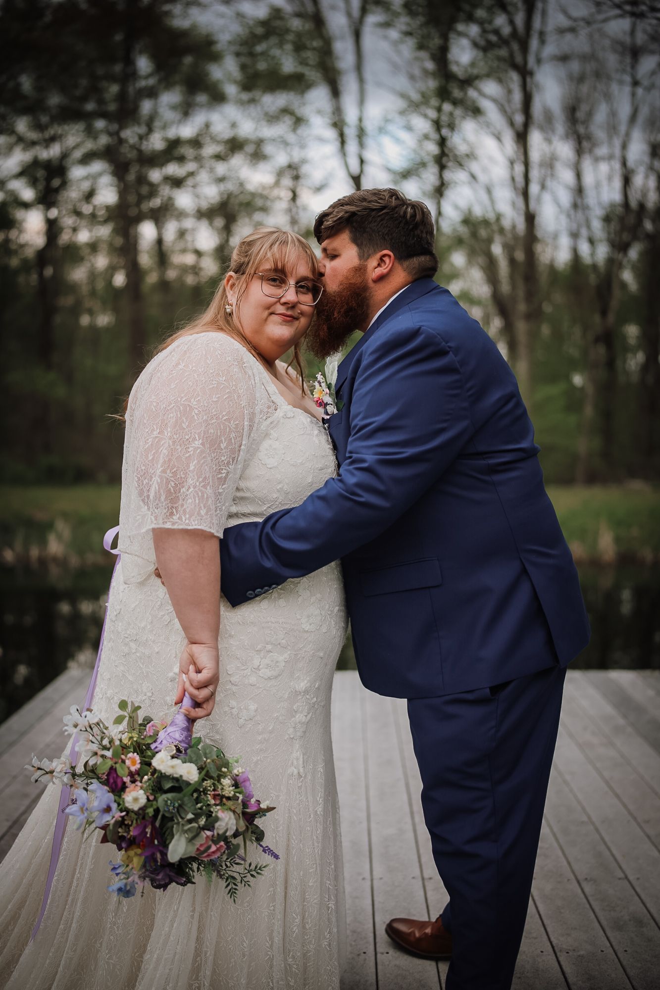 A groom in a blue suit kisses his bride on the cheek as she holds a purple floral bouquet on a wooden dock.