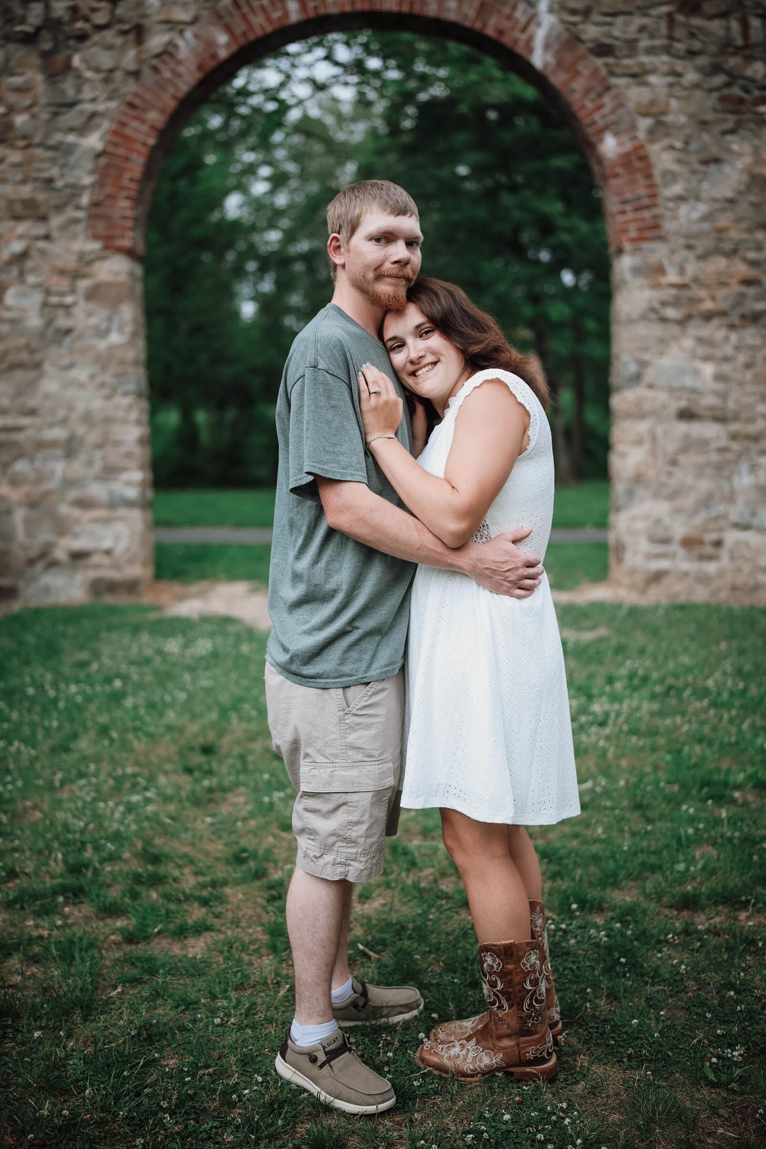 Couple embracing outdoors near a brick archway, woman in white dress and boots, man in gray shirt and khaki shorts.