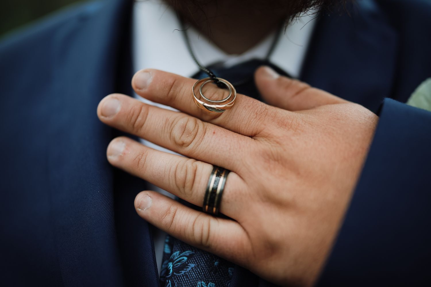 Close-up of a groom's hand wearing a black band ring, holding two gold wedding rings against a blue suit.