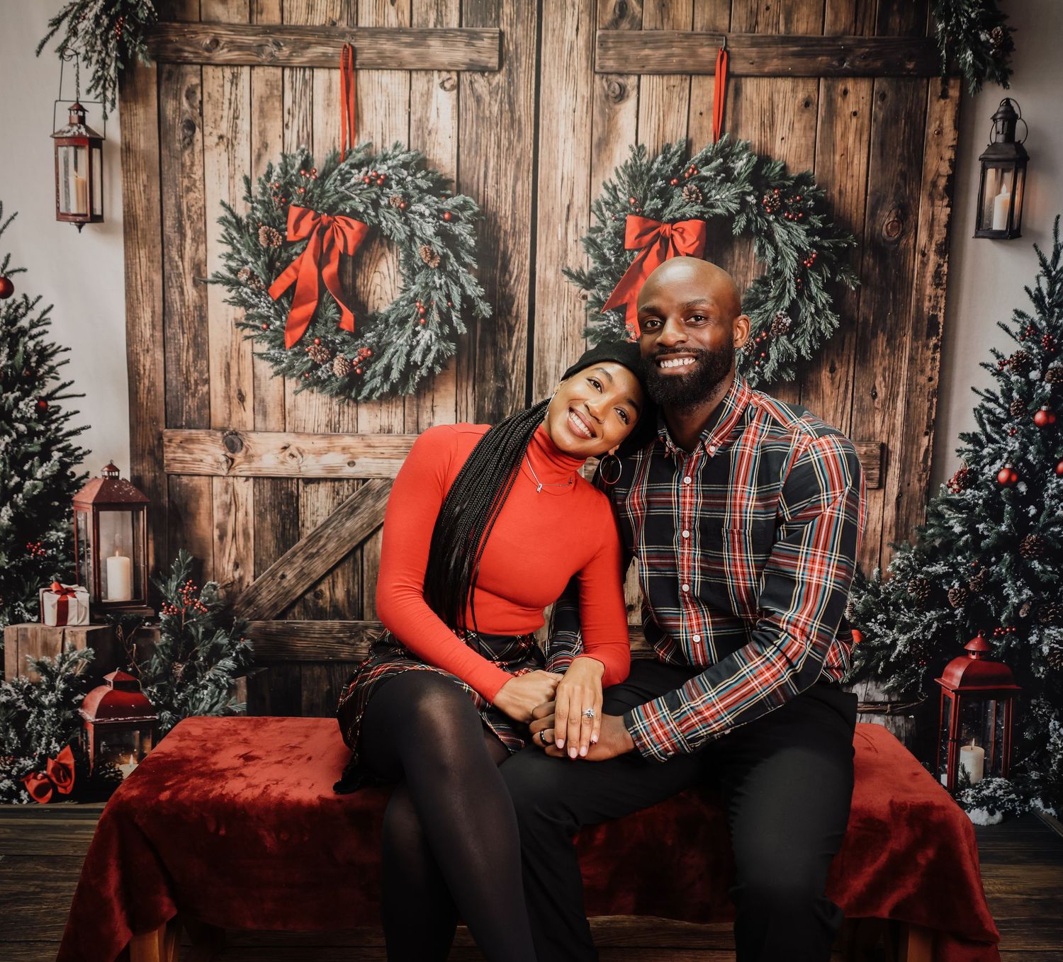 Couple smiling together in front of rustic wooden doors with Christmas wreaths and holiday decorations.