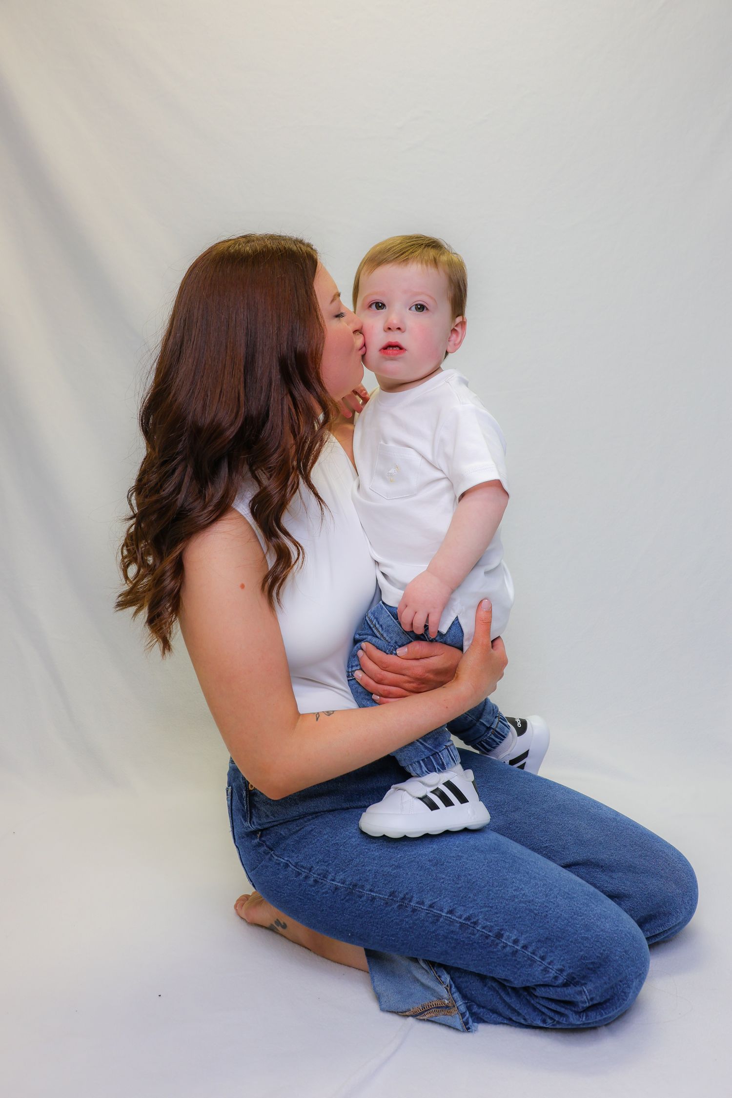A mother kneeling on white backdrop kisses her baby boy in white tee and jeans, both wearing matching outfits.