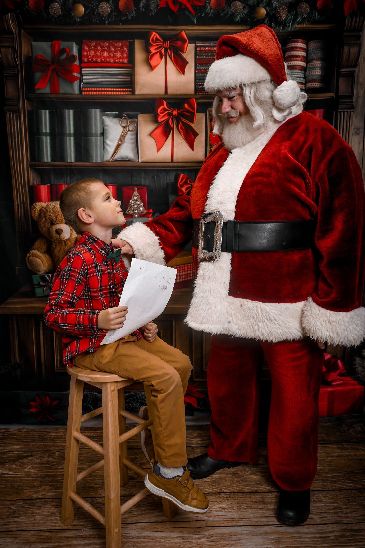 A boy in plaid shirt reads his Christmas wish list to Santa Claus in a festive decorated room with gifts.