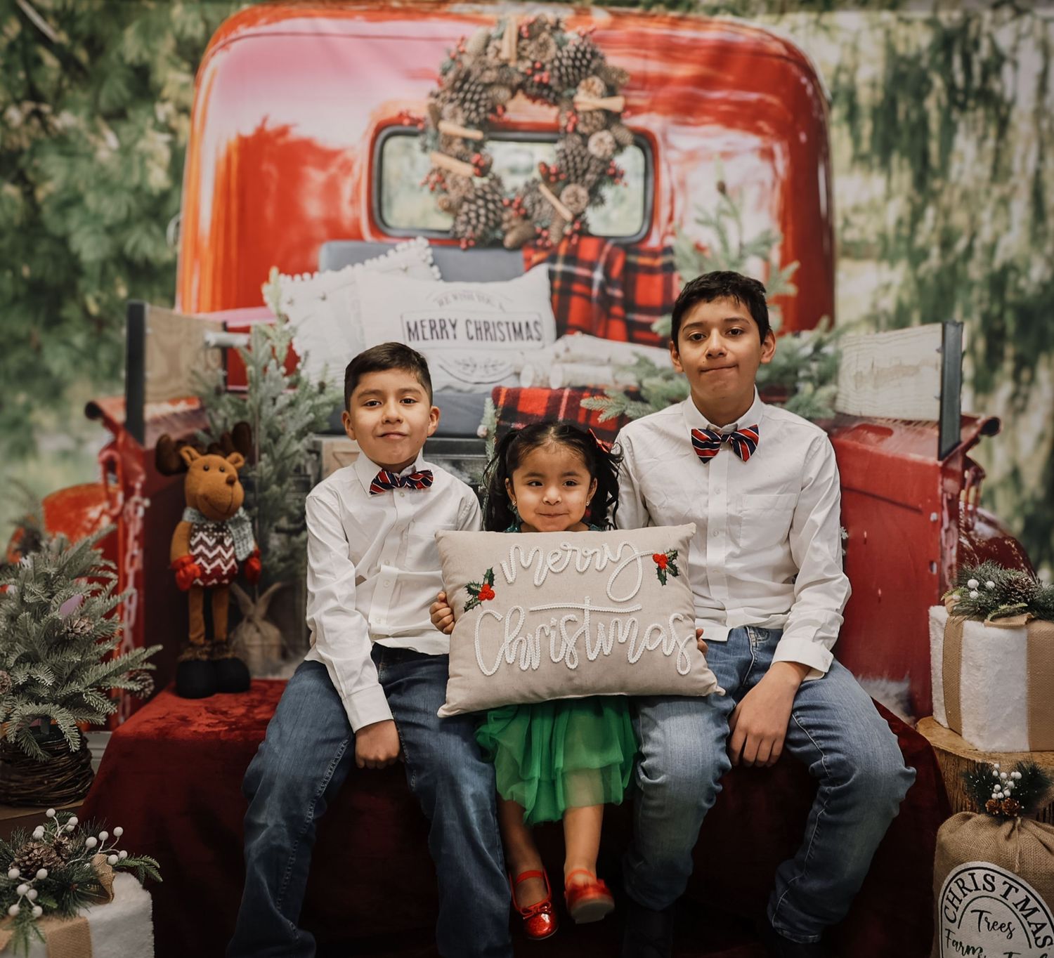 Three children pose for a festive Christmas photo with a red truck backdrop, holding a Merry Christmas pillow.