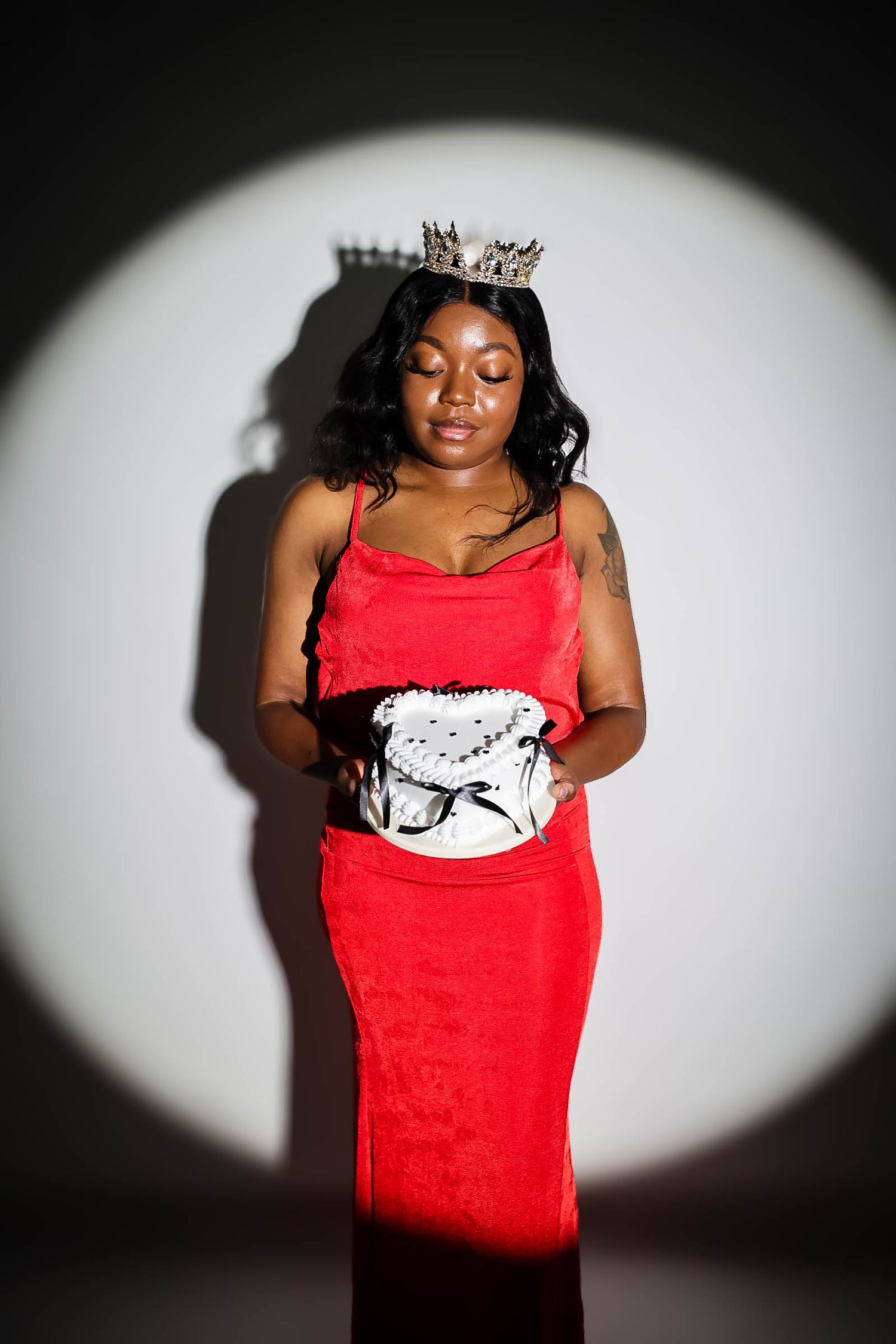 Woman in red velvet dress and crown poses under spotlight against white background.