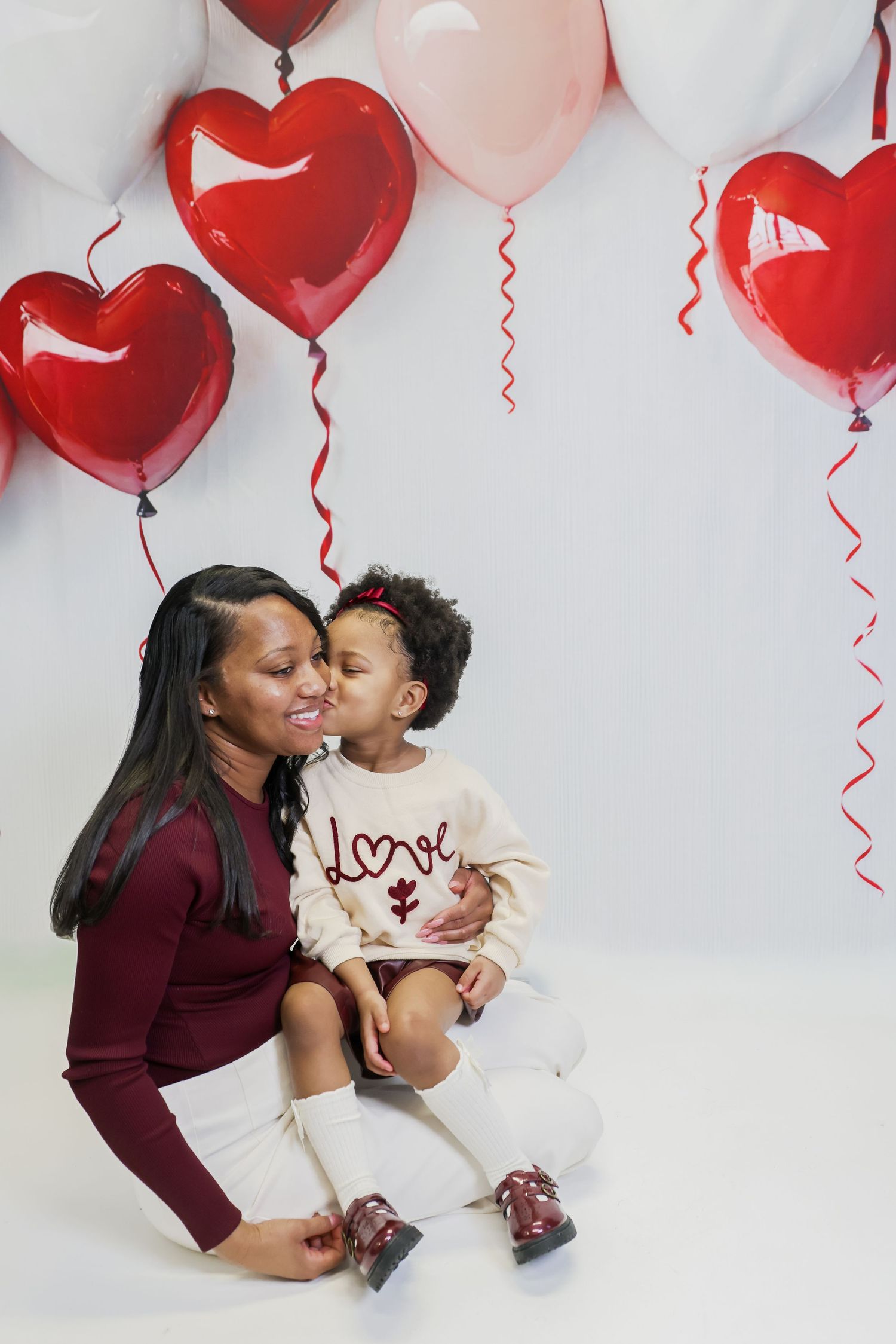 A mother and toddler share a sweet kiss surrounded by red and pink heart balloons on Valentine's Day.