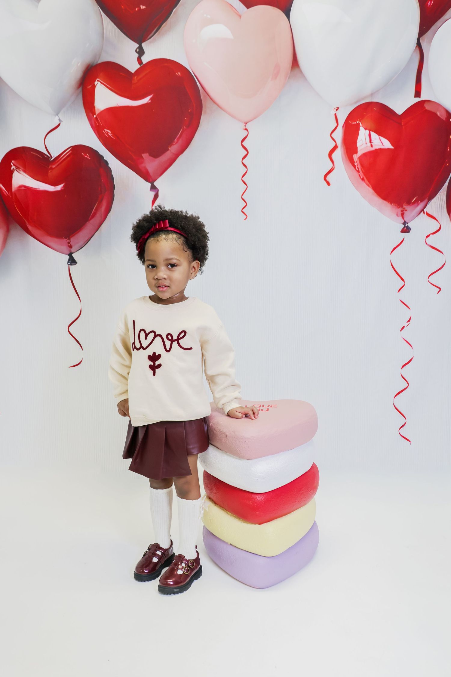 Young girl in a love sweatshirt and burgundy skirt poses with heart-shaped boxes and Valentine's Day balloons.