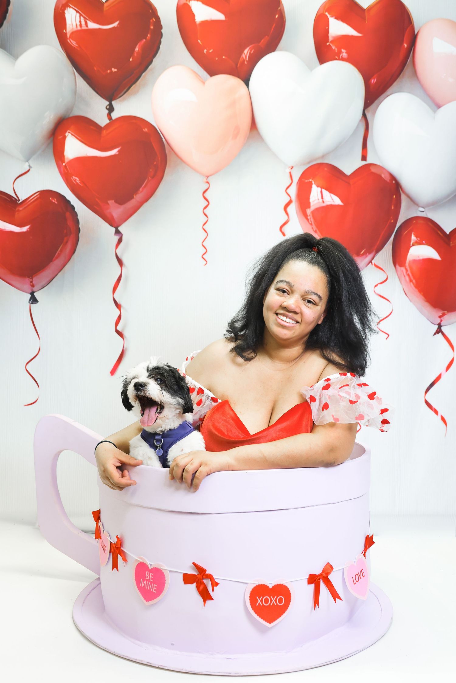 Woman smiling in giant teacup with dog, surrounded by red and white heart balloons for Valentine's Day.