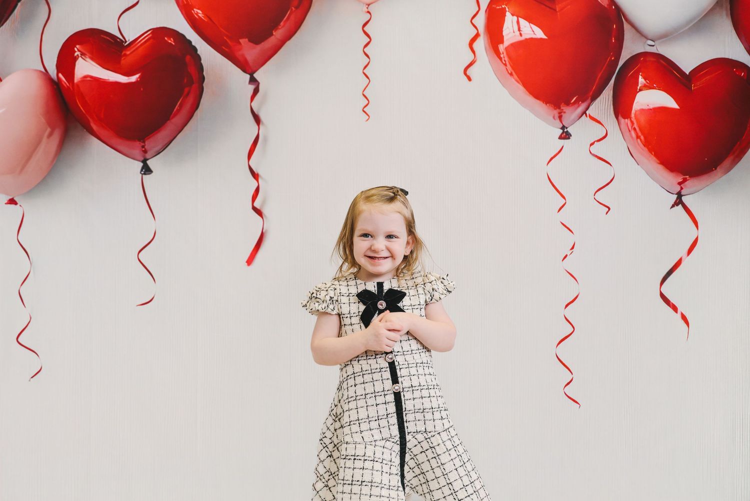 A smiling little girl in a plaid dress holds a bow while standing in front of red heart-shaped balloons on a white wall.