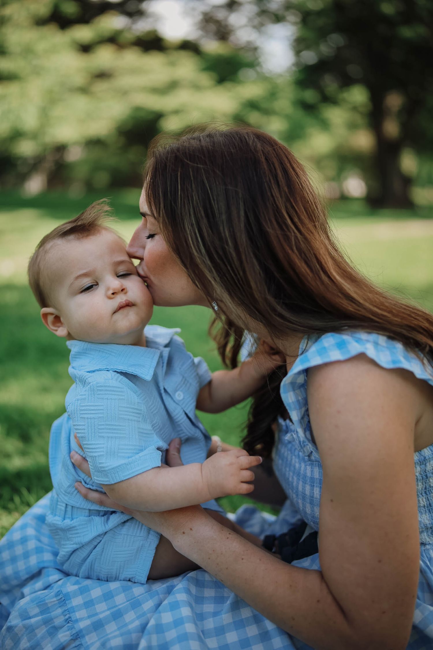 A mother in a blue gingham dress kisses her baby boy outdoors in a lush green park setting.