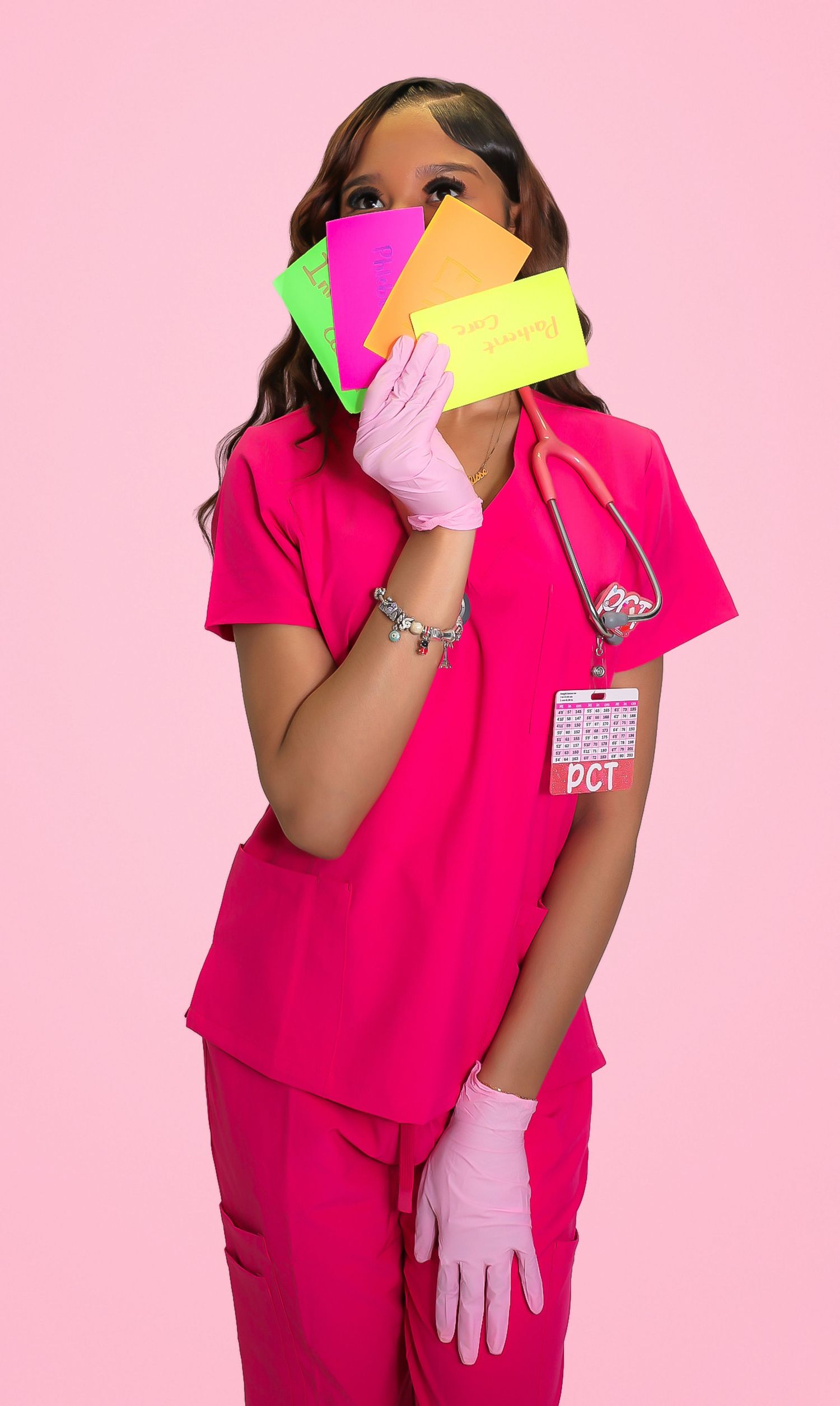 A female PCT in pink scrubs holds colorful sticky notes, wearing gloves and stethoscope against a pink background.