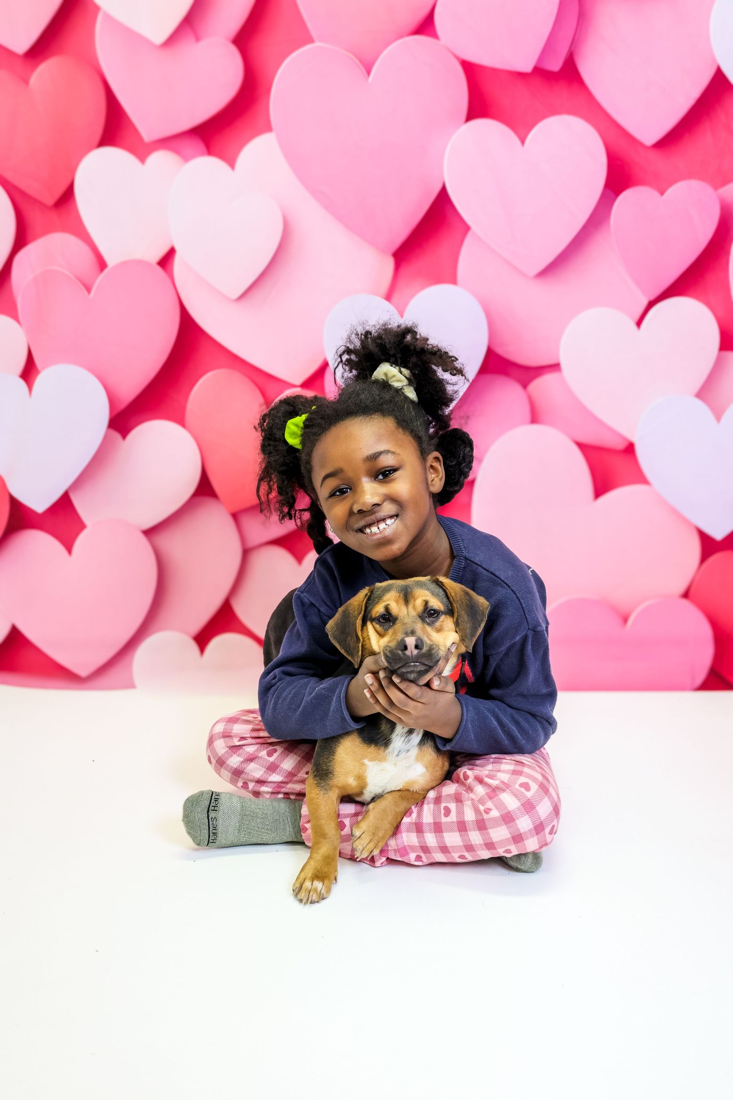 A young child lies on the floor with a small brown puppy against a pink heart-patterned backdrop.