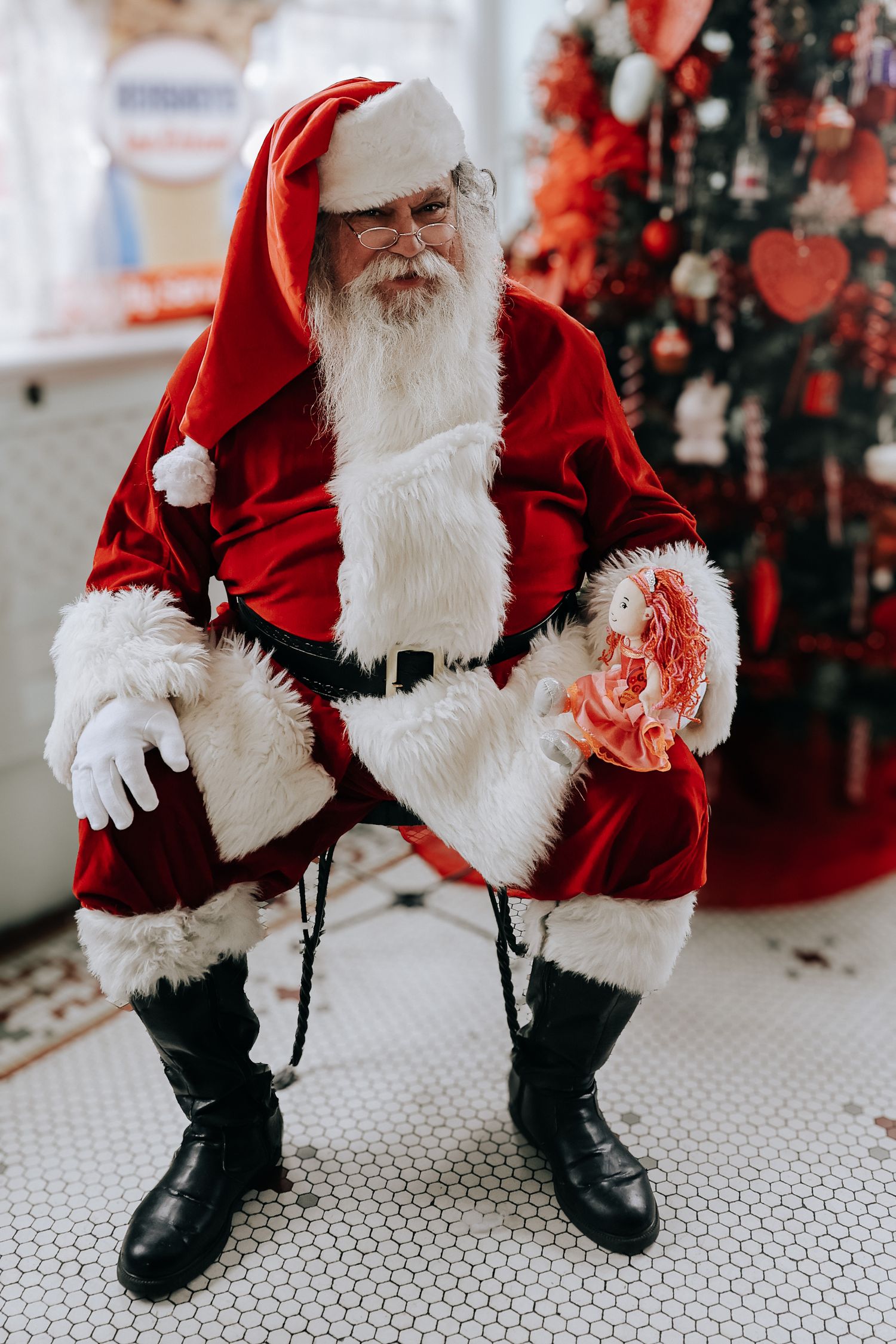 Santa Claus in traditional red suit sitting on hexagonal floor tiles with Christmas decorations in background.