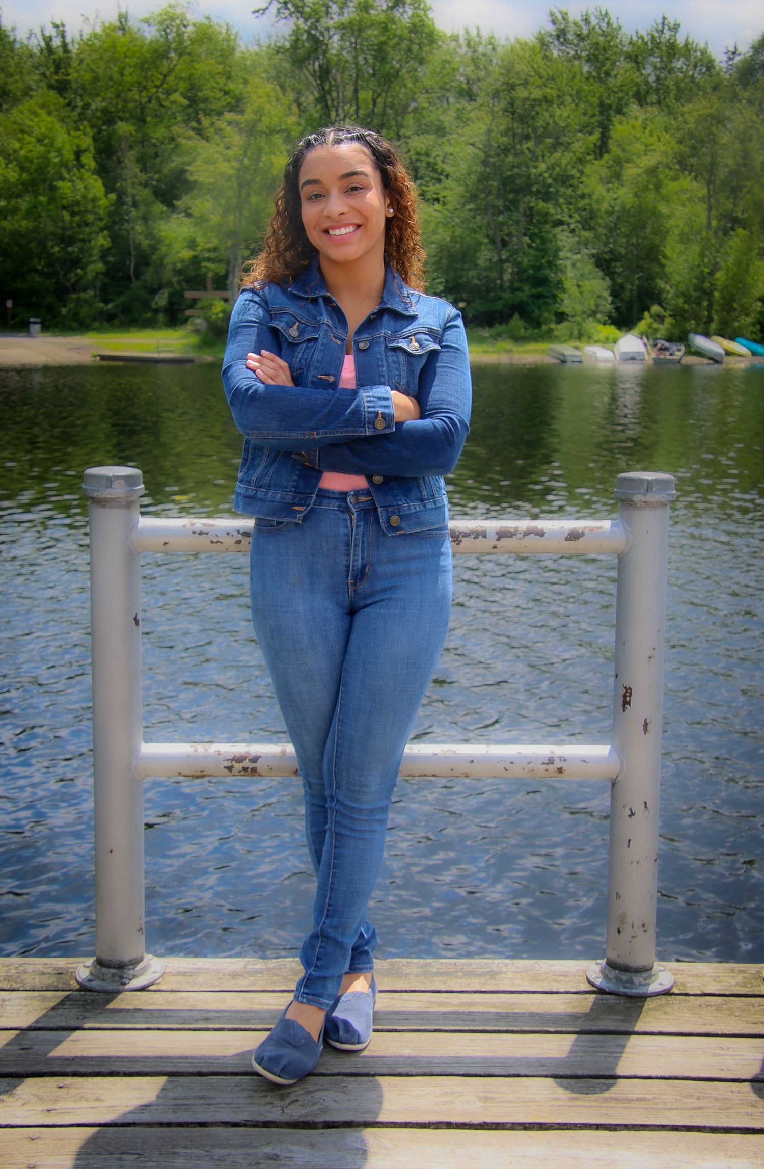 Person in denim outfit stands on wooden dock overlooking peaceful lake surrounded by green trees.